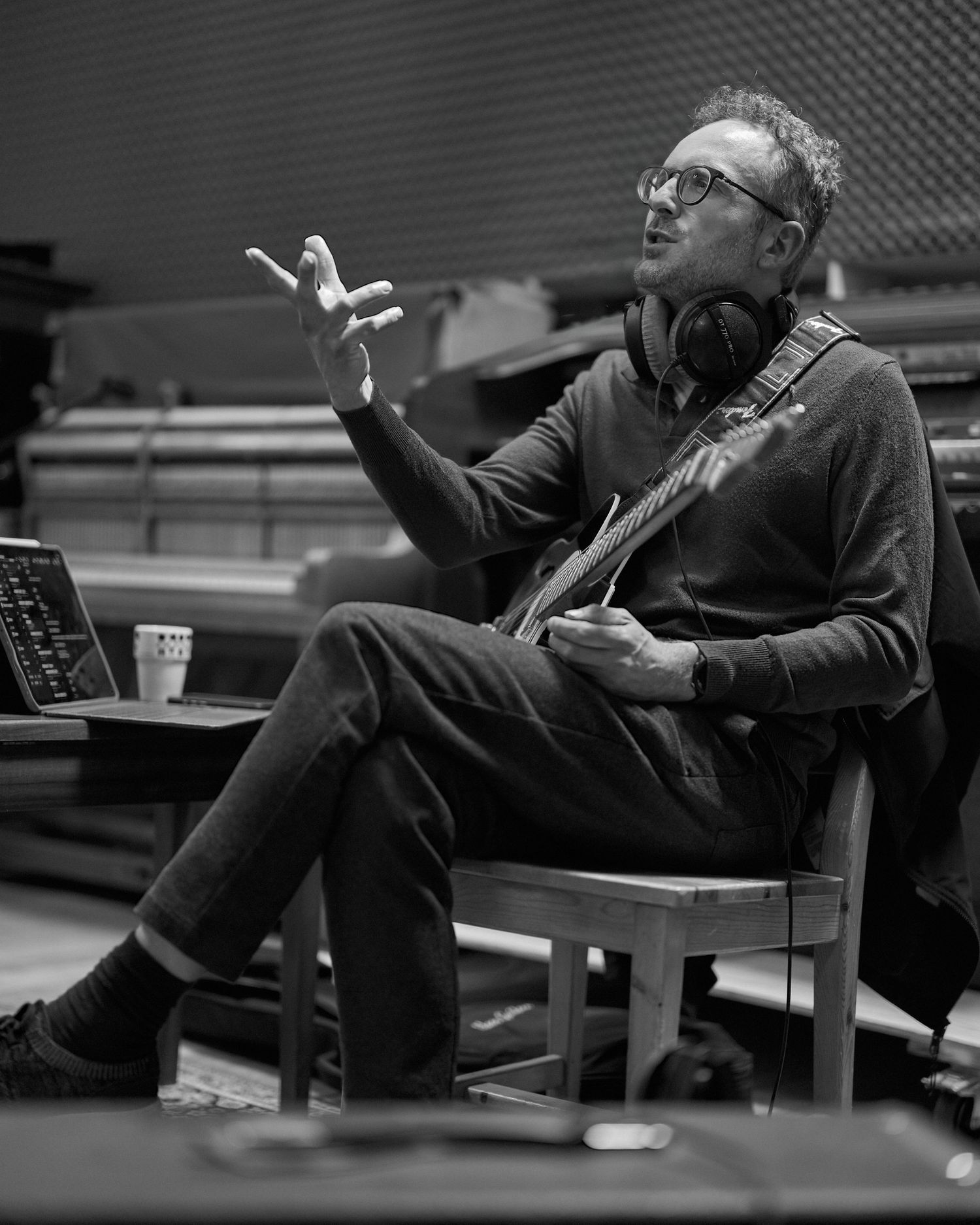 Black and white photo of a man with headphones around his neck, sitting and gesturing while holding a guitar.