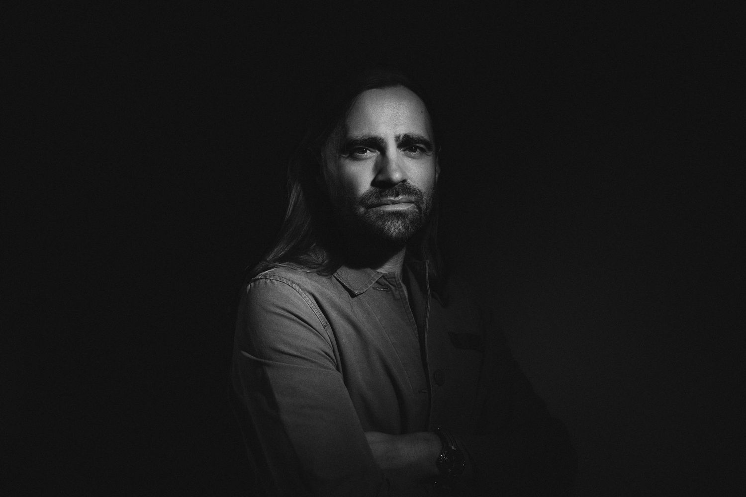 Black and white portrait of a bearded man with long hair, arms crossed, against a dark background.
