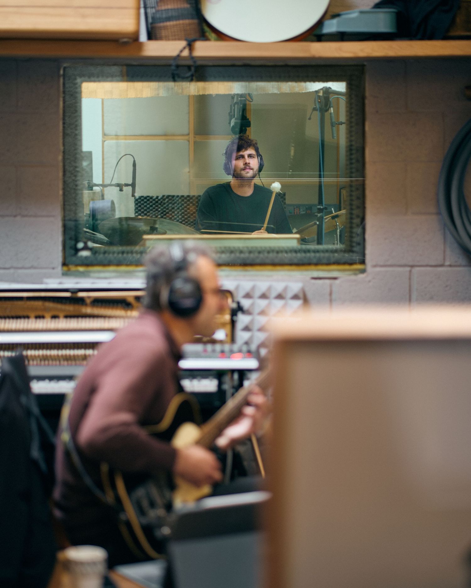 Two musicians recording in a studio, one playing guitar with headphones, another visible through a glass window behind a microphone.
