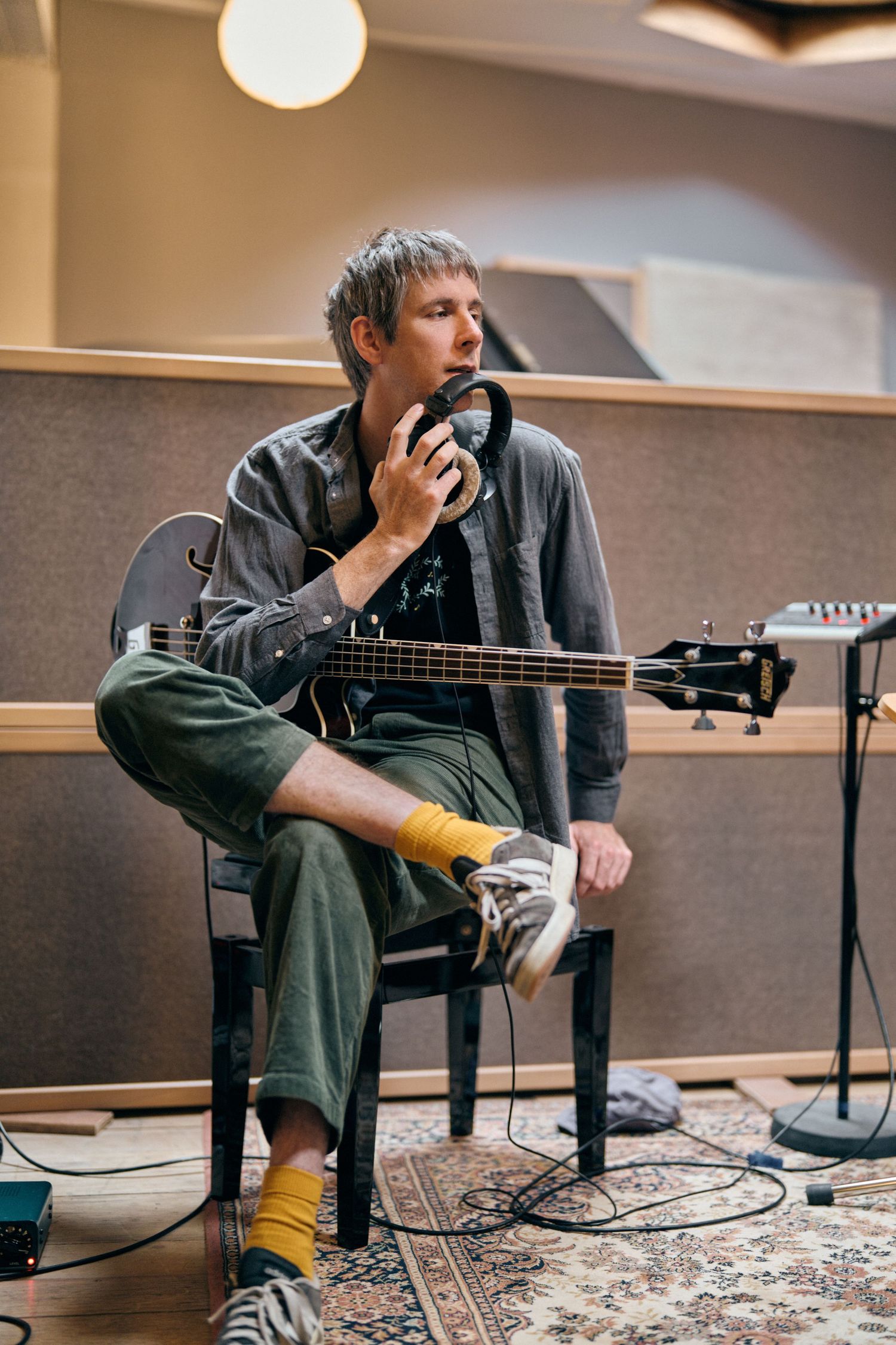 Musician sitting in recording studio holding headphones while playing bass guitar, wearing green pants and yellow socks.
