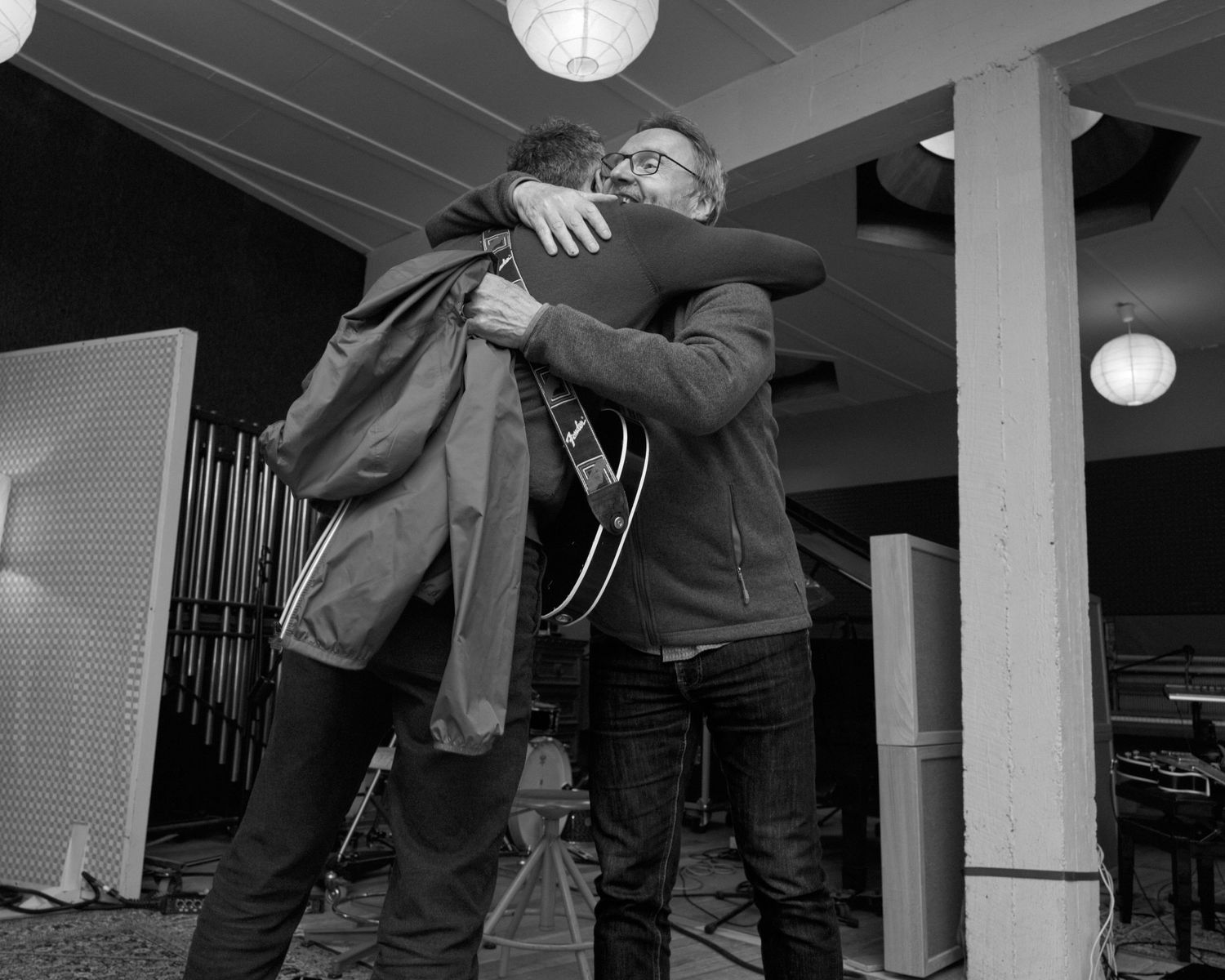 Two men share a warm hug indoors in a black and white photo, with a guitar visible in the background.