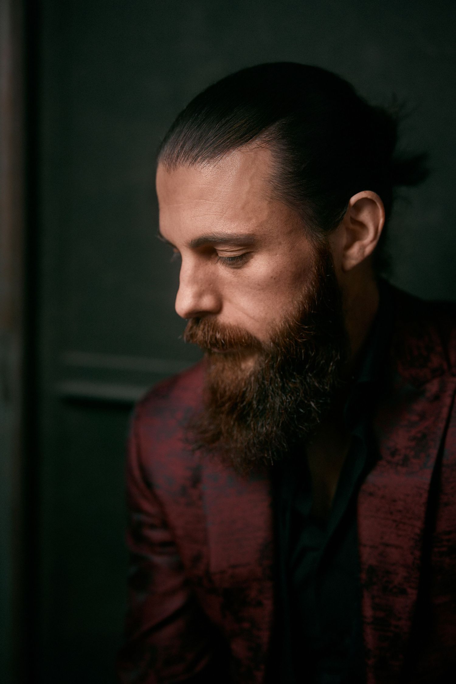 Bearded man with hair in bun wearing dark red patterned blazer, looking down in moody dark green setting.