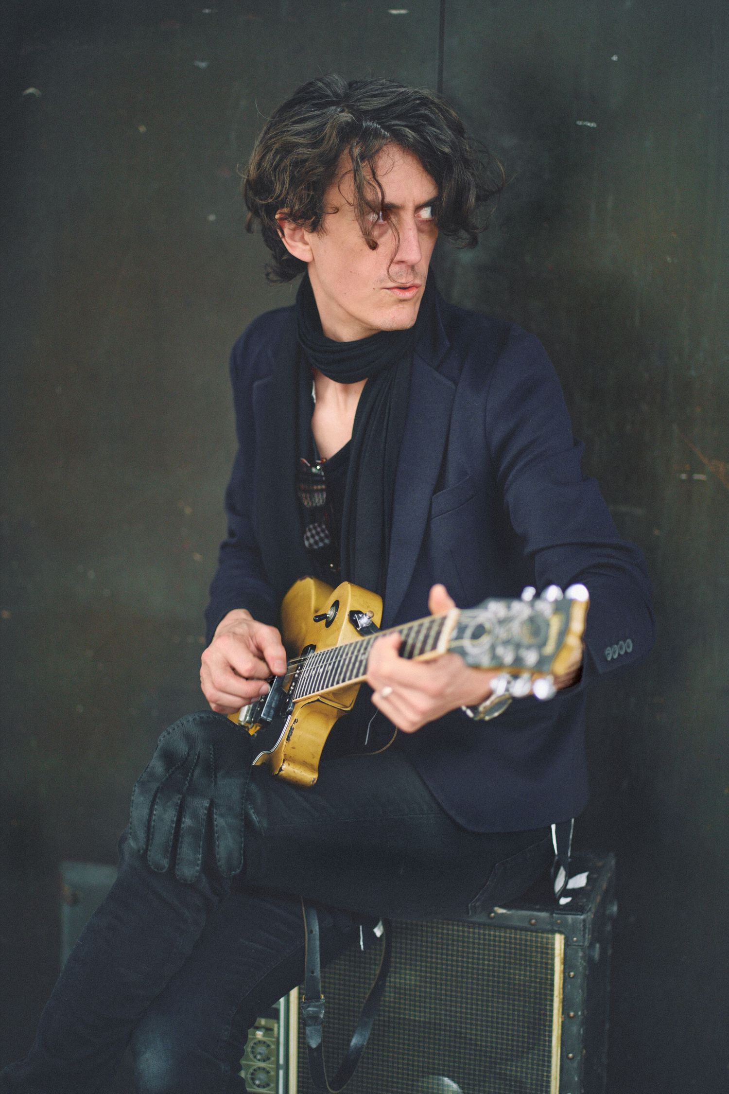 Curly-haired male guitarist in a dark blazer playing a gold electric guitar while seated on an amp.
