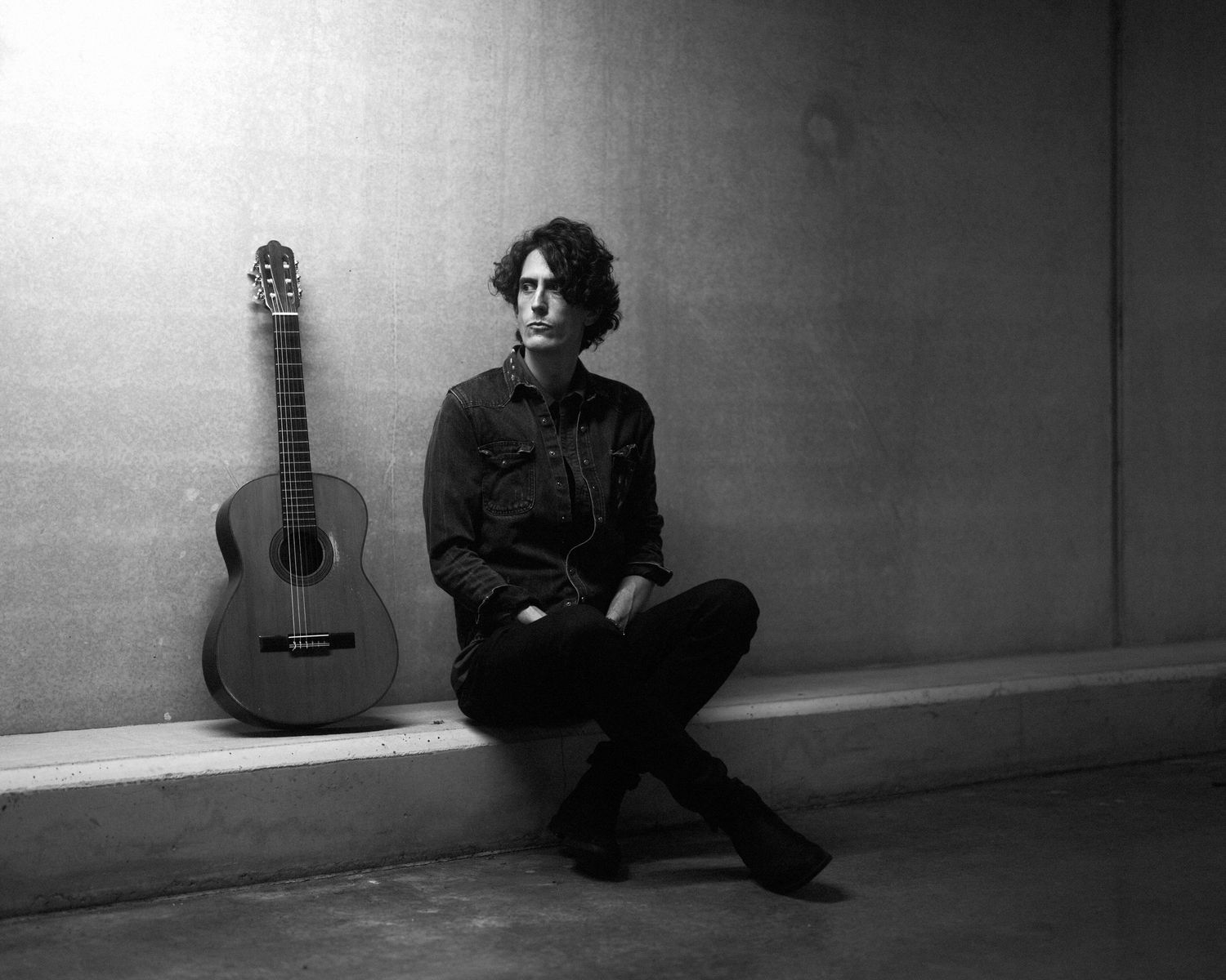 Young man seated beside acoustic guitar against concrete wall in black and white portrait.
