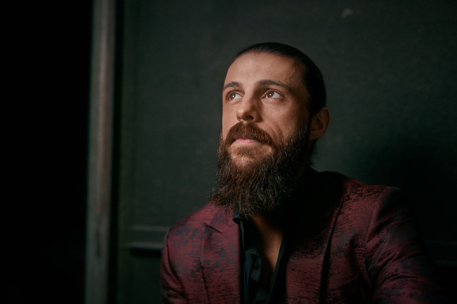Bearded man in a red patterned blazer gazing upward thoughtfully against a dark background.