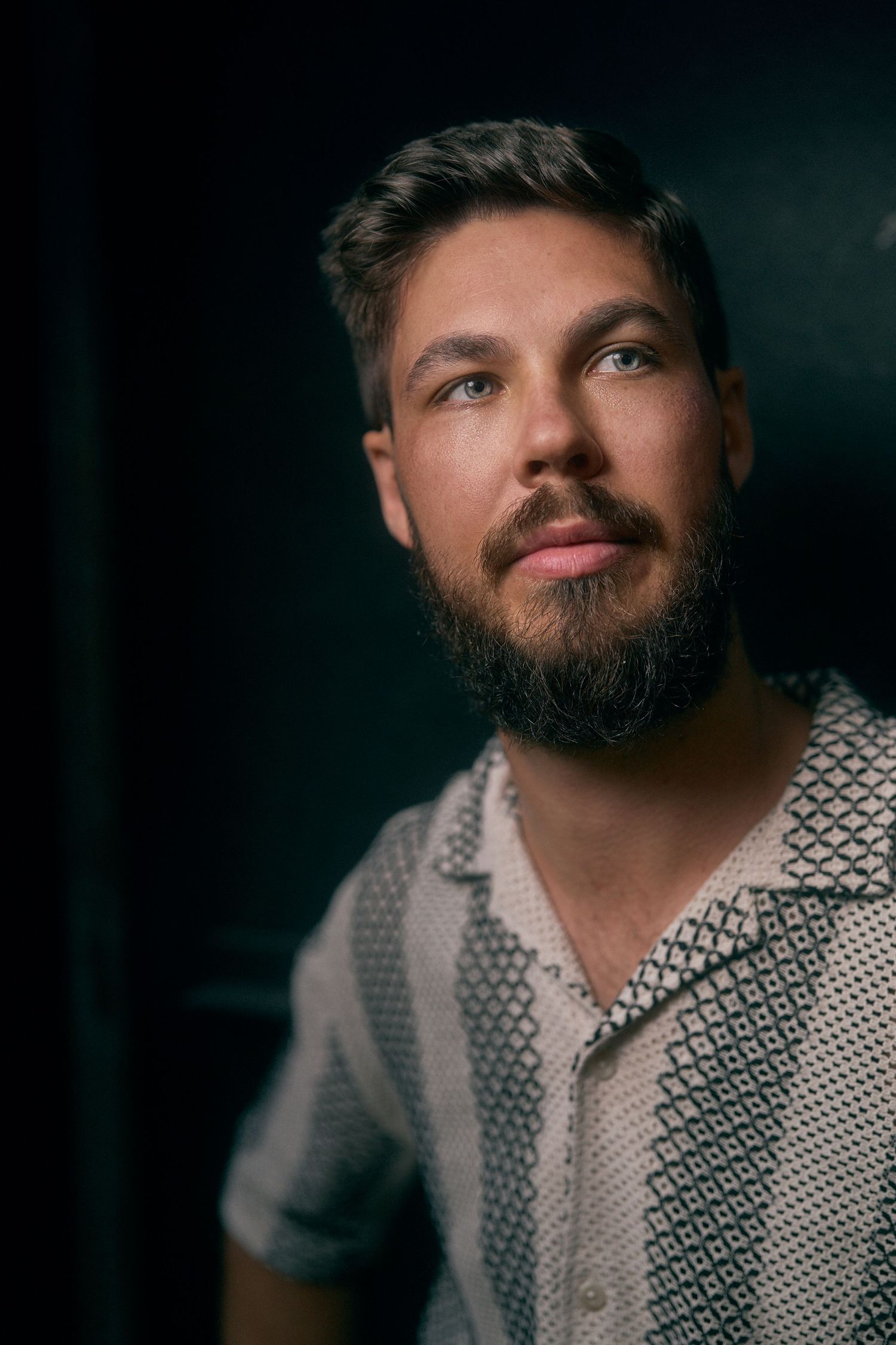 Bearded man in patterned polo shirt posing for portrait against dark background with soft lighting.