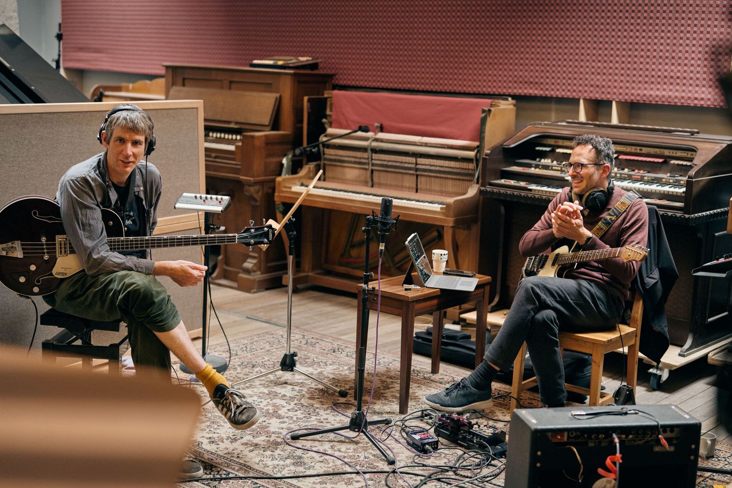 Two musicians recording in a professional studio, one with headphones playing bass, the other strumming guitar.