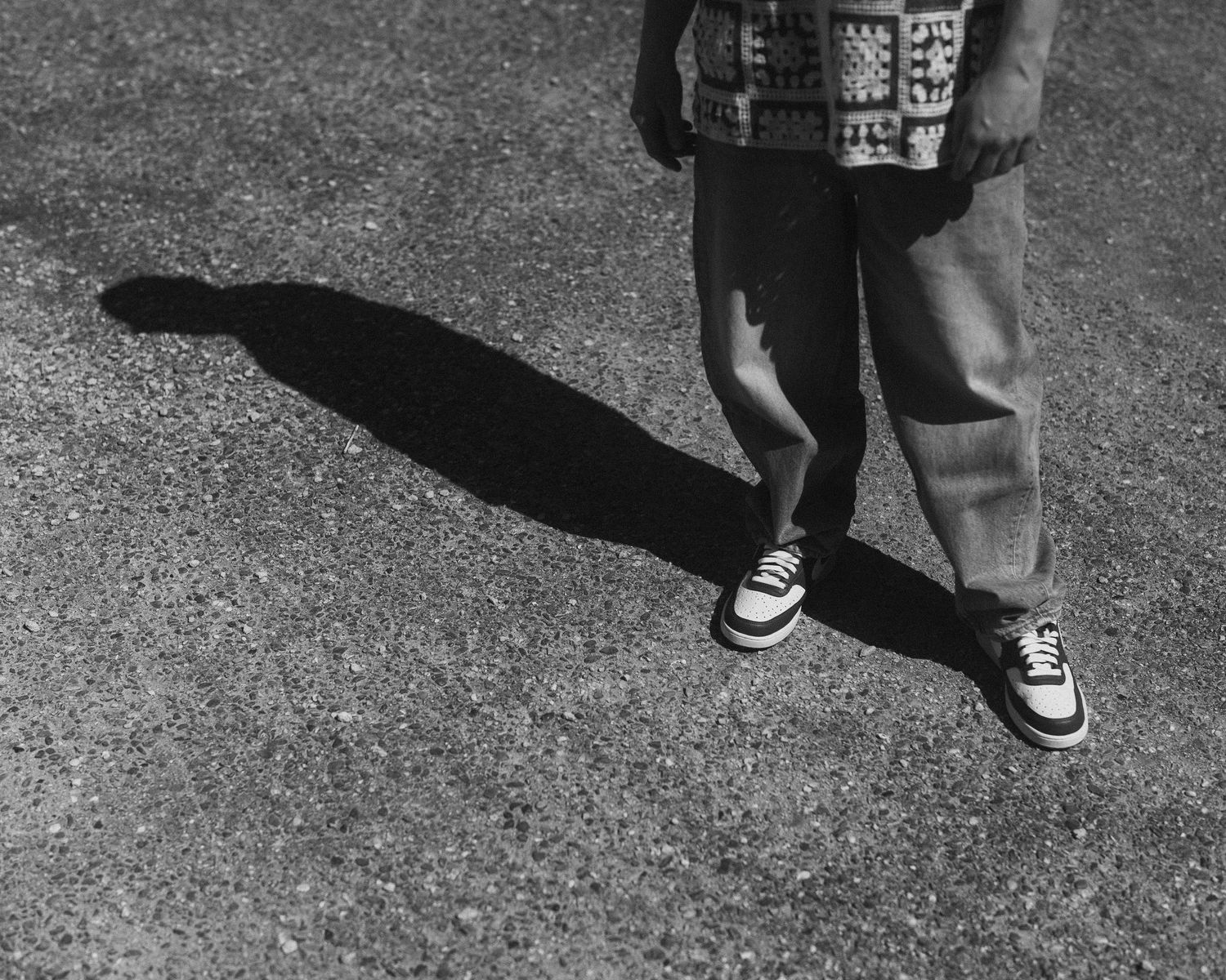 Black and white photo of a person standing on pavement casting a long shadow wearing Vans sneakers.