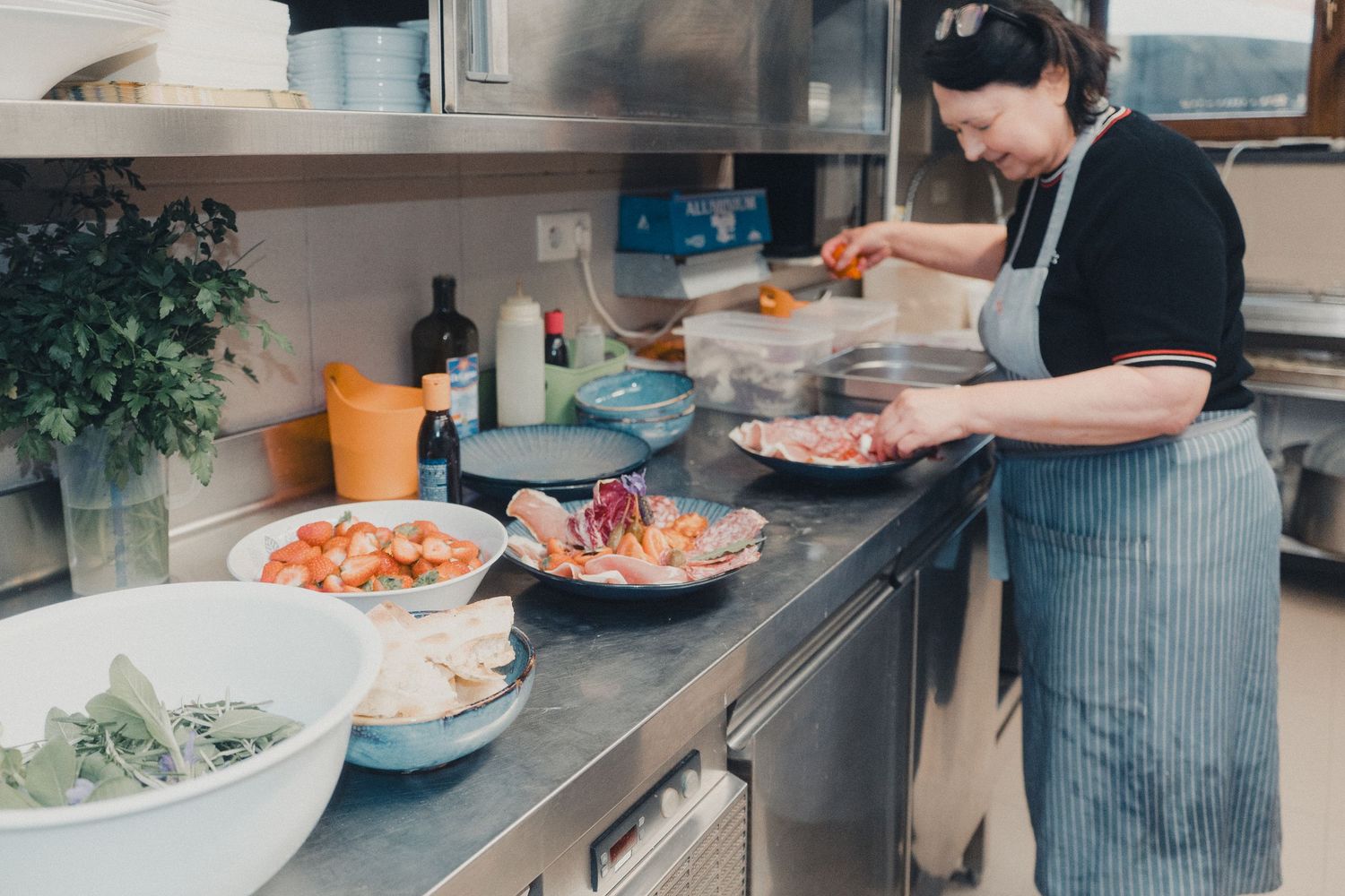 A woman in an apron prepares fresh ingredients in a professional kitchen surrounded by bowls of vegetables.