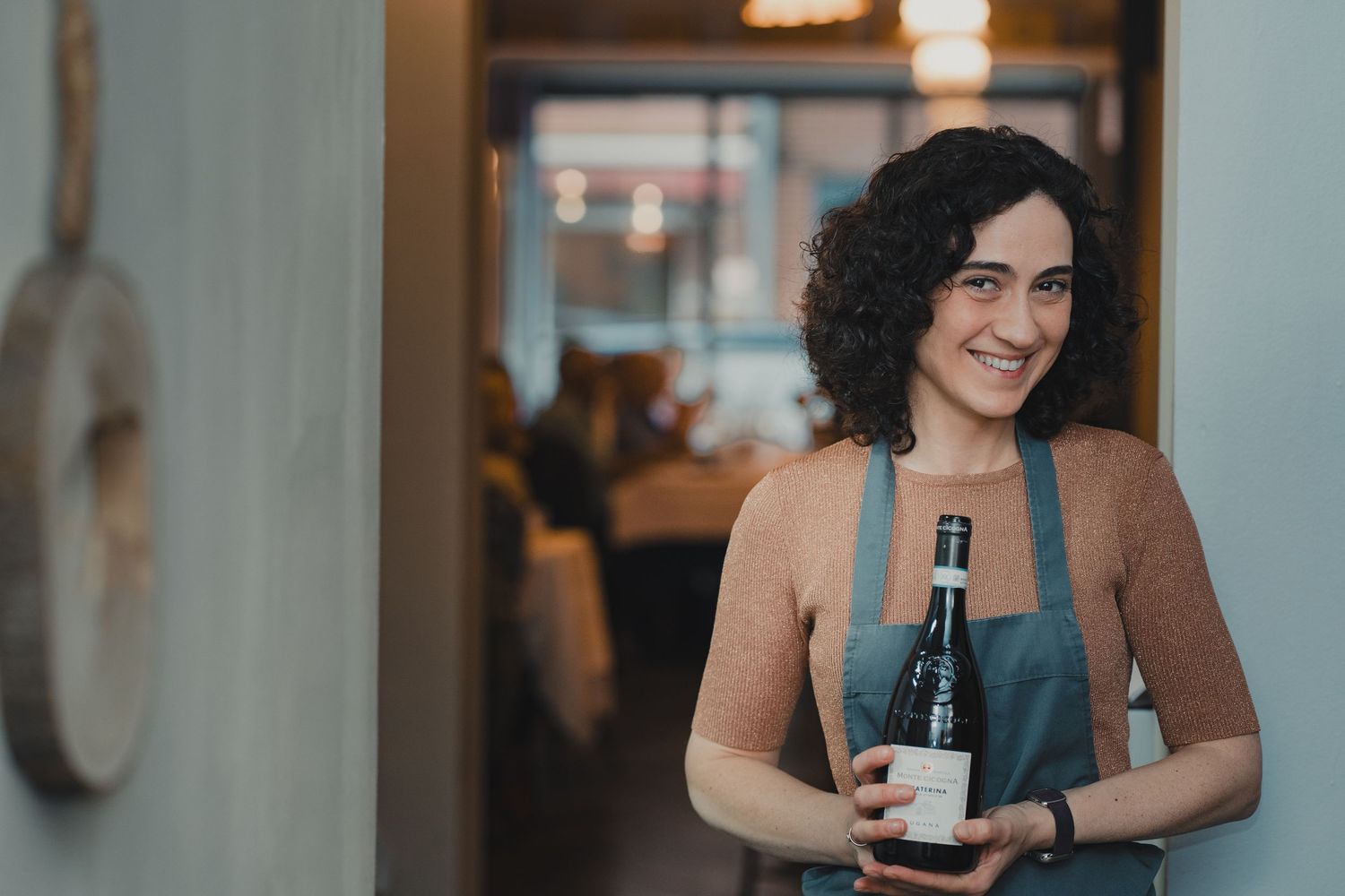 Smiling waitress in apron holding a wine bottle in a restaurant setting.