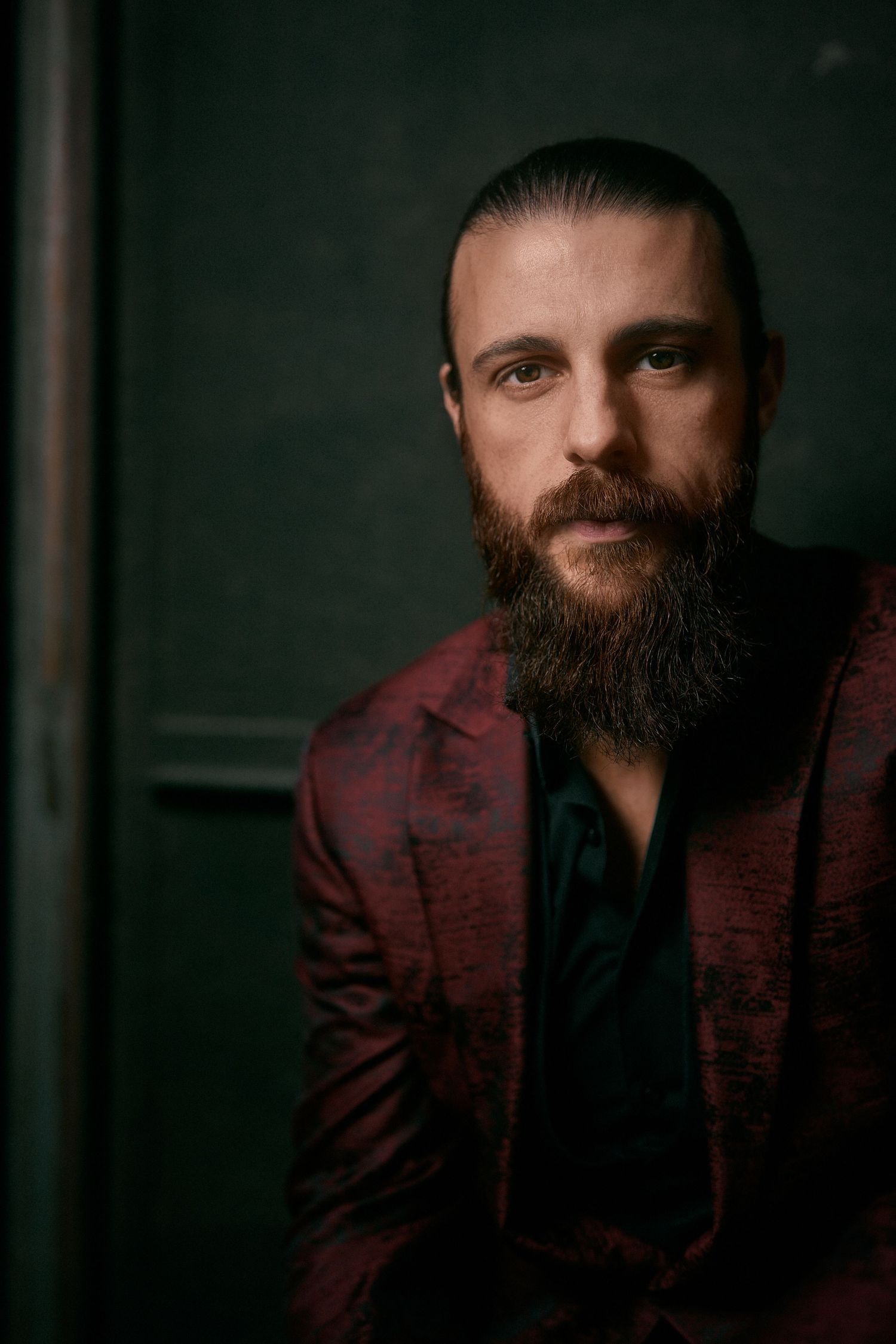 Bearded man wearing a red patterned blazer in a dramatic dark green studio portrait.