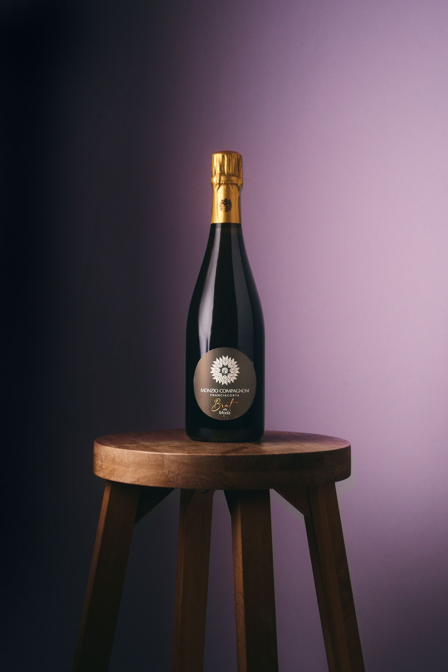 Elegant champagne bottle with gold foil top displayed on a rustic wooden stool against a purple gradient background.