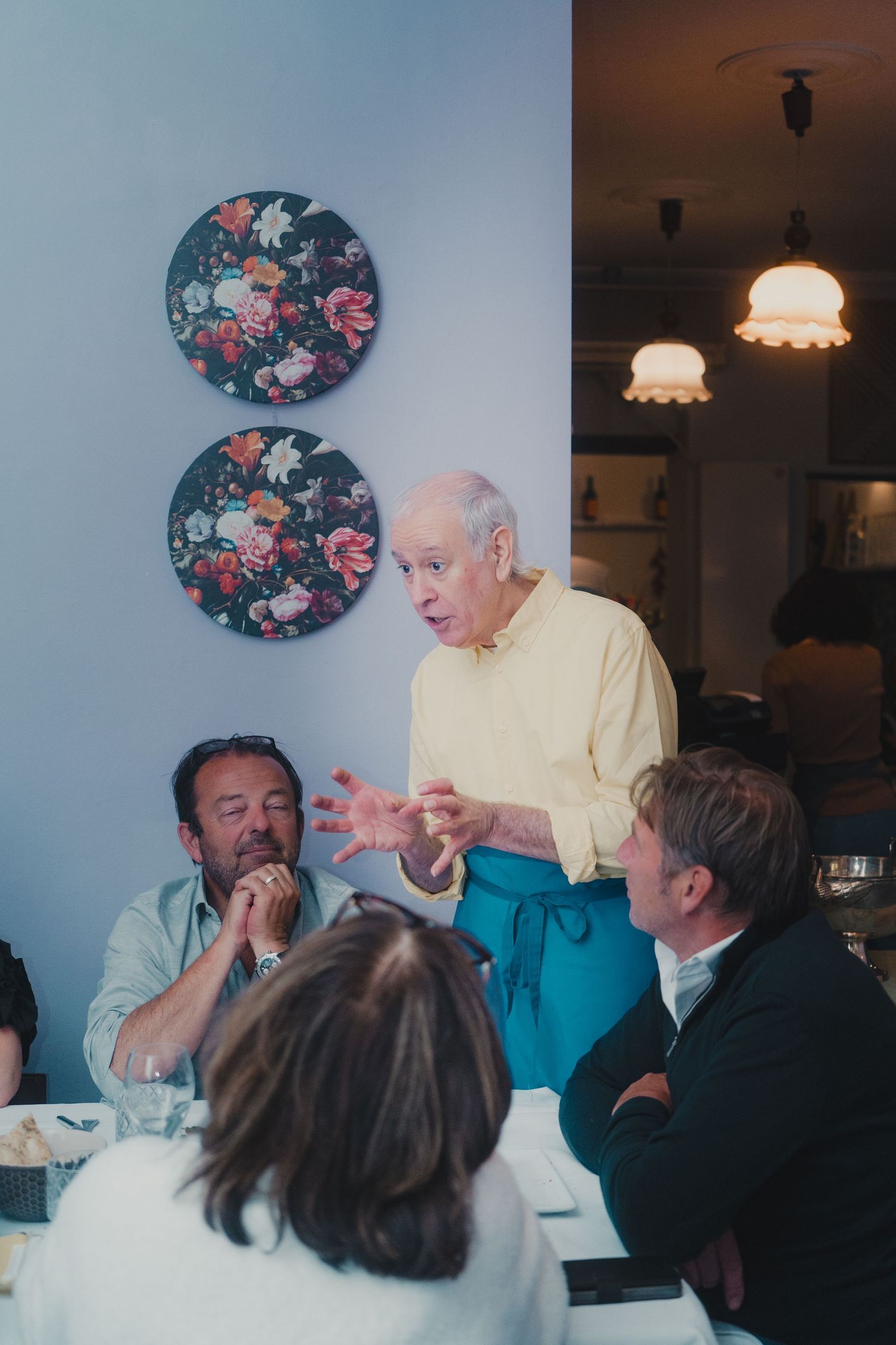 Elderly man in yellow shirt and blue apron speaks to seated diners in a restaurant with floral wall decor.