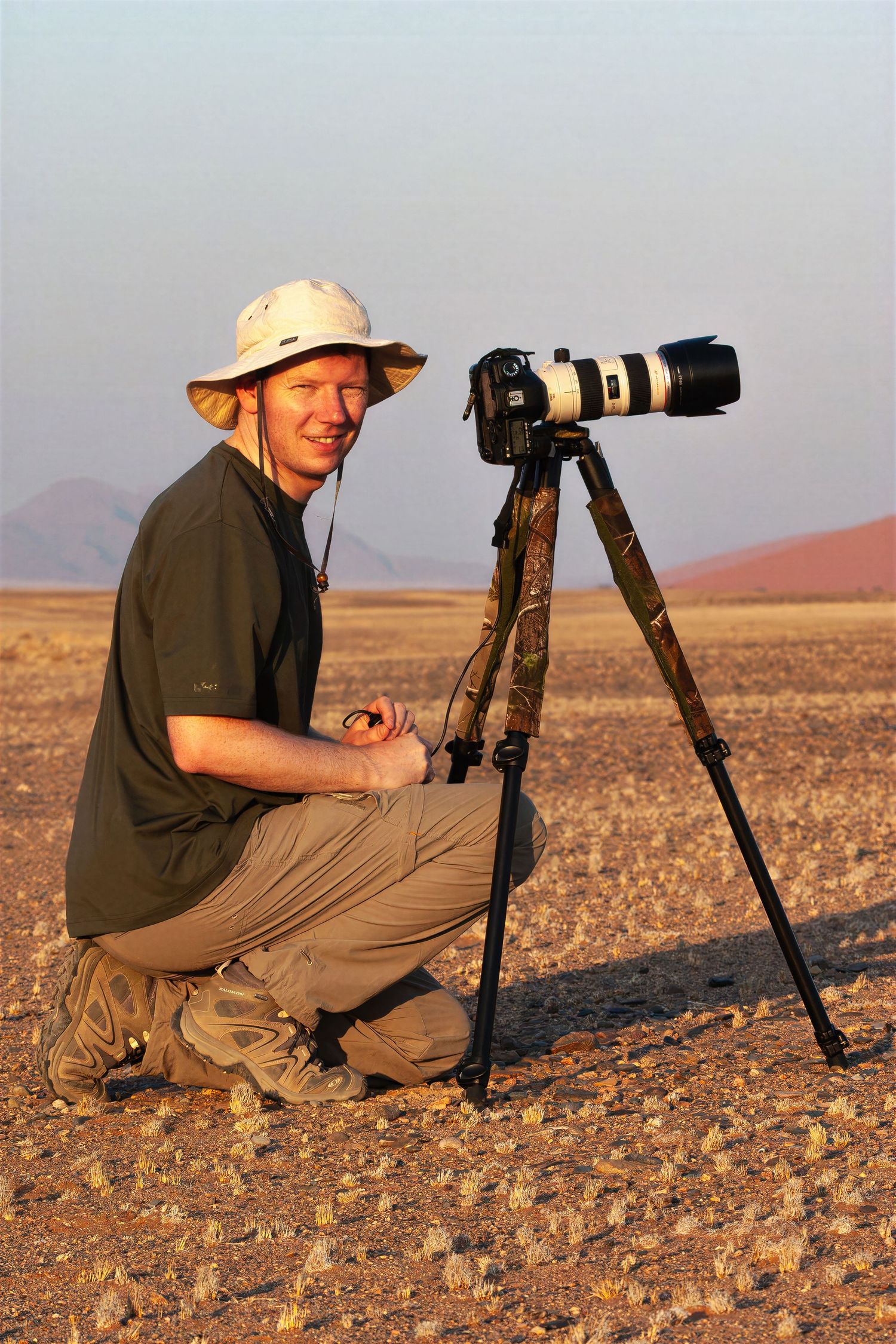 A wildlife photographer crouches beside a camera on a tripod in the Namib Desert at golden hour.