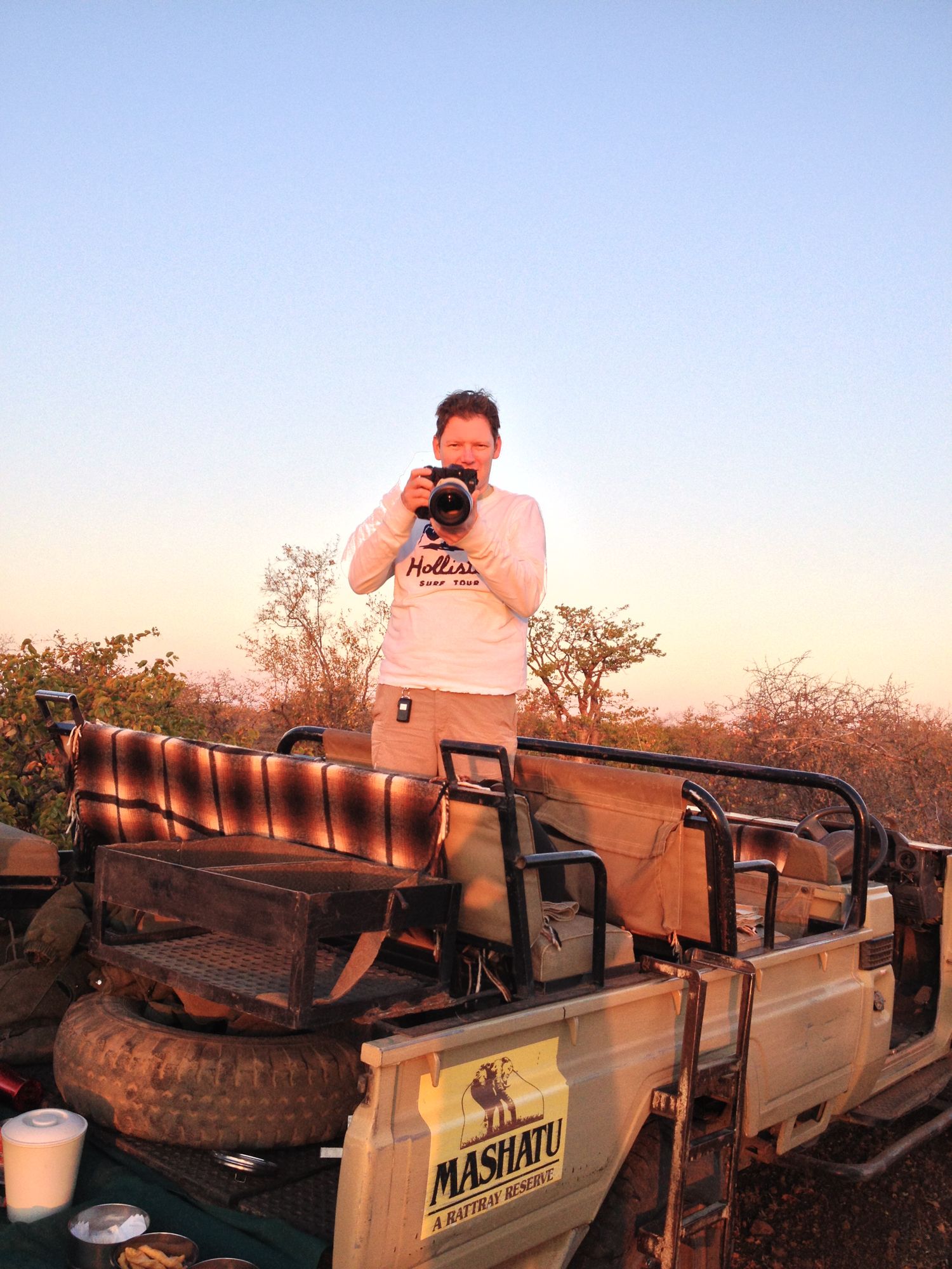 A person holding a camera on a safari vehicle in the African bush.