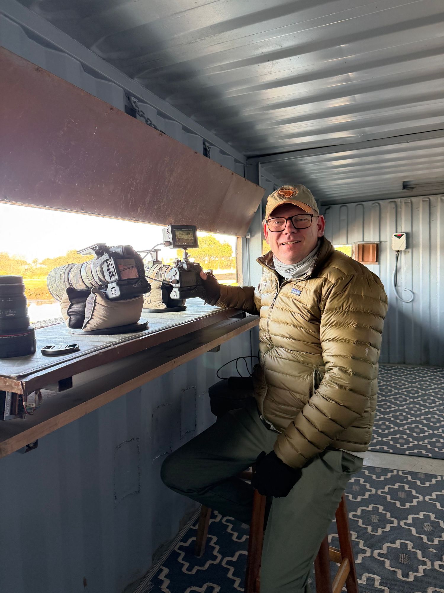Man smiling inside a photography hide with camera equipment on a shelf in warm morning light.