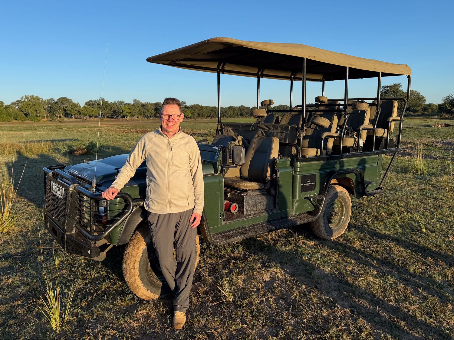 Man posing next to a green open-sided safari game drive vehicle on an African savanna at golden hour.