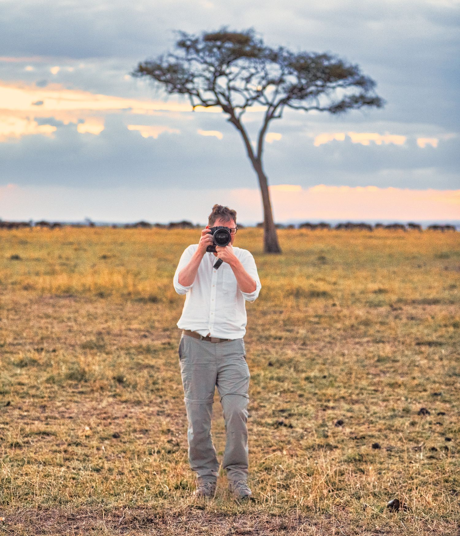 A wildlife photographer aims his camera on the open African savanna with an acacia tree behind.