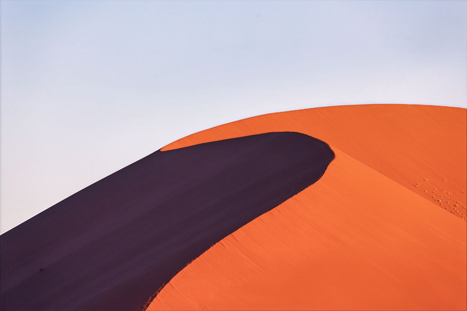 A dramatic orange Namib Desert sand dune with a sharp ridge casting deep shadows under a pale sky.