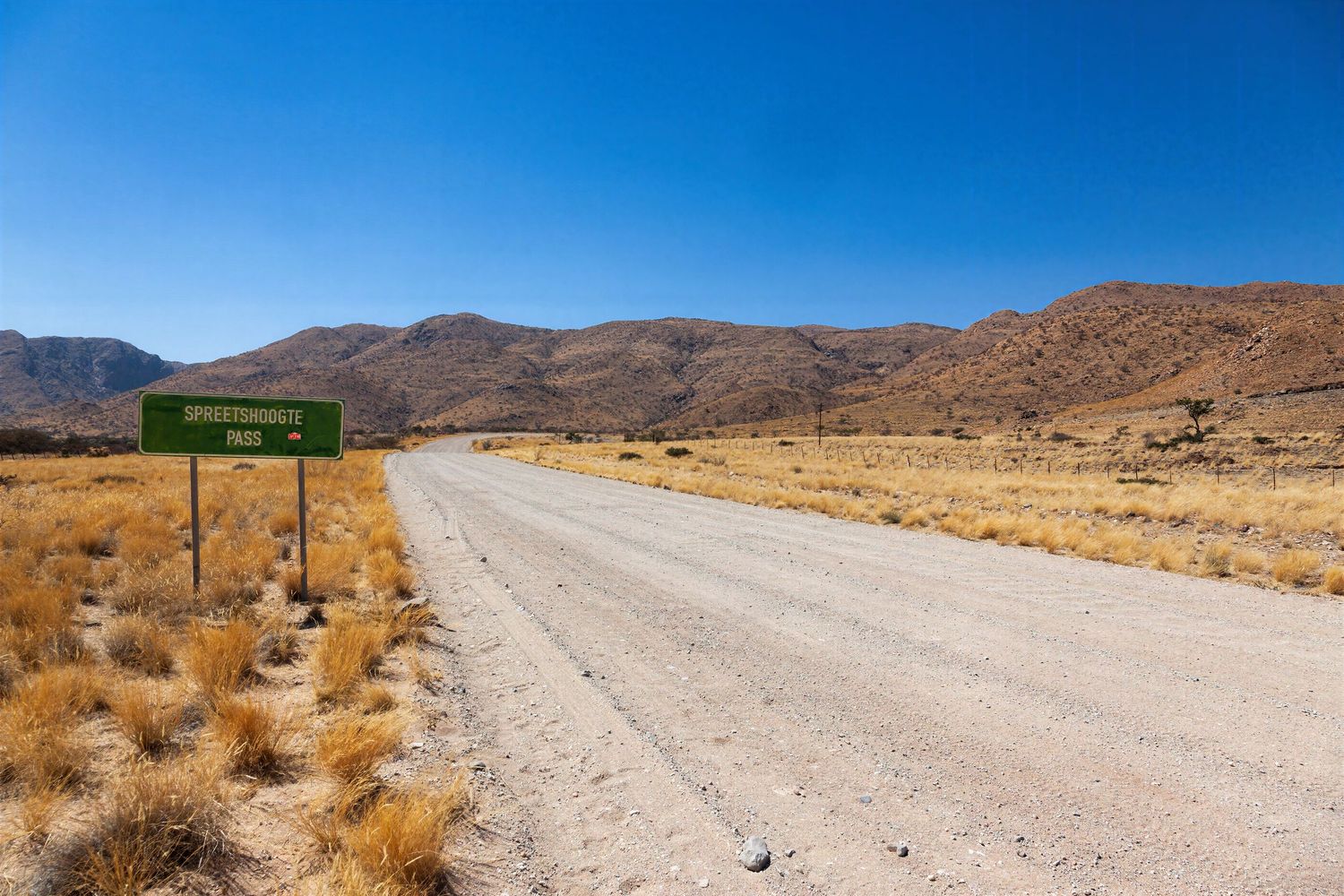 A gravel road leads through arid Namibian landscape with dry grass and rocky hills under a clear blue sky.
