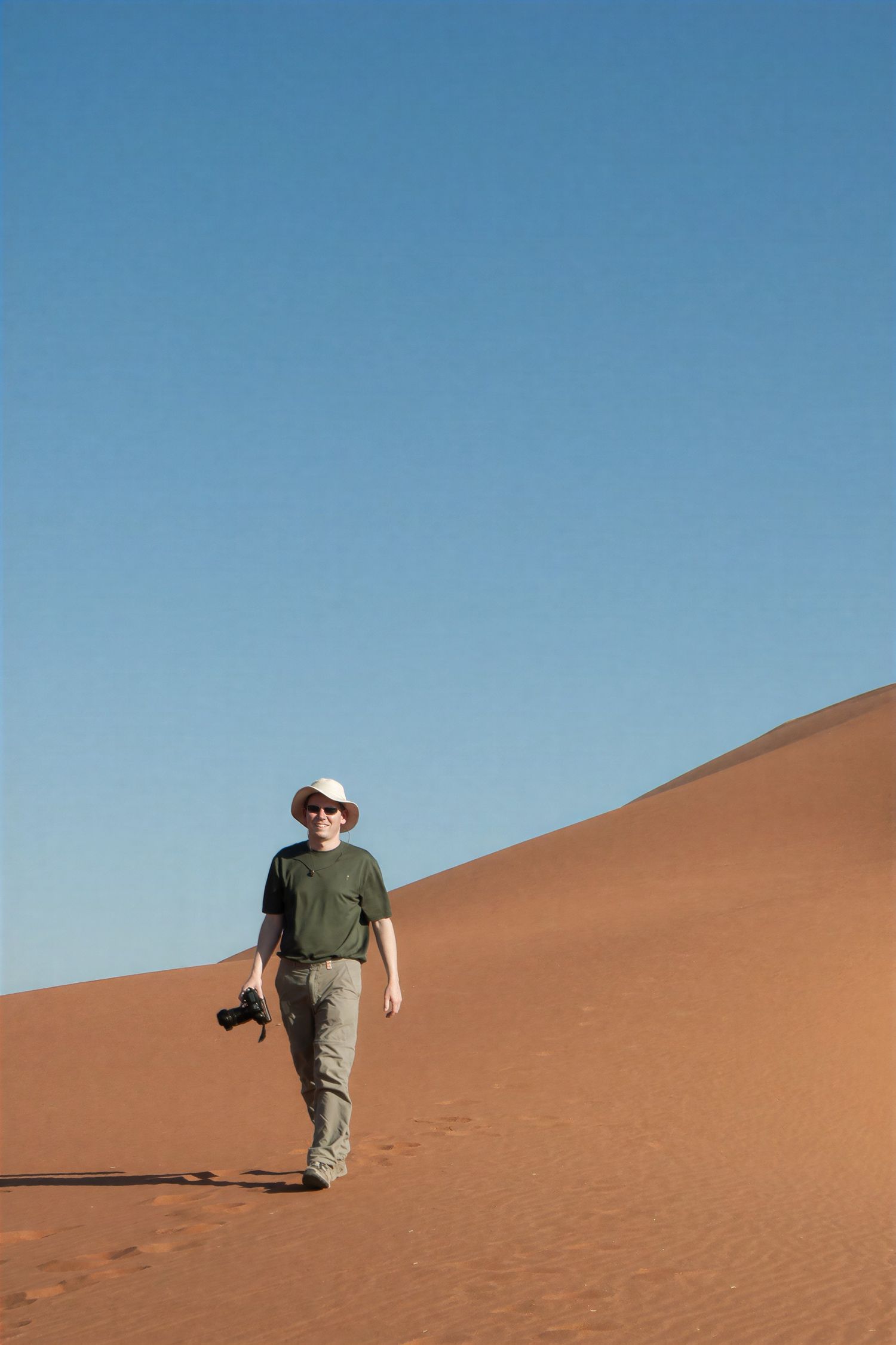 A photographer walks across a vast red sand dune under a clear blue sky in a desert landscape.