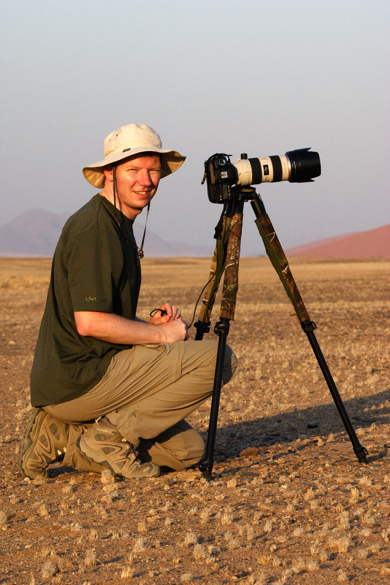 Photographer kneeling beside a DSLR camera with telephoto lens on tripod in a vast desert landscape.