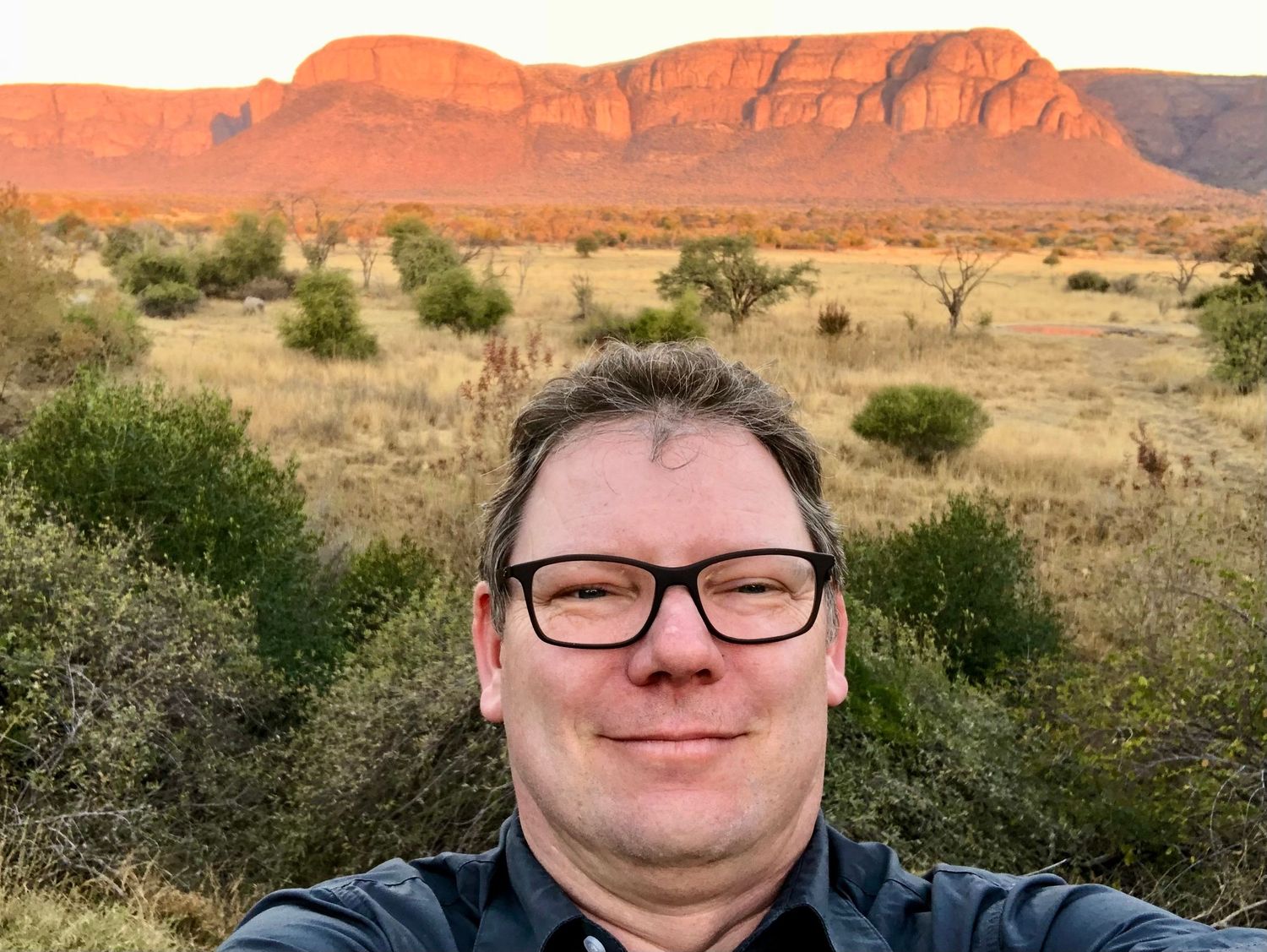 Man taking a selfie at sunset with dramatic red rocky mountain landscape glowing behind him in Africa.