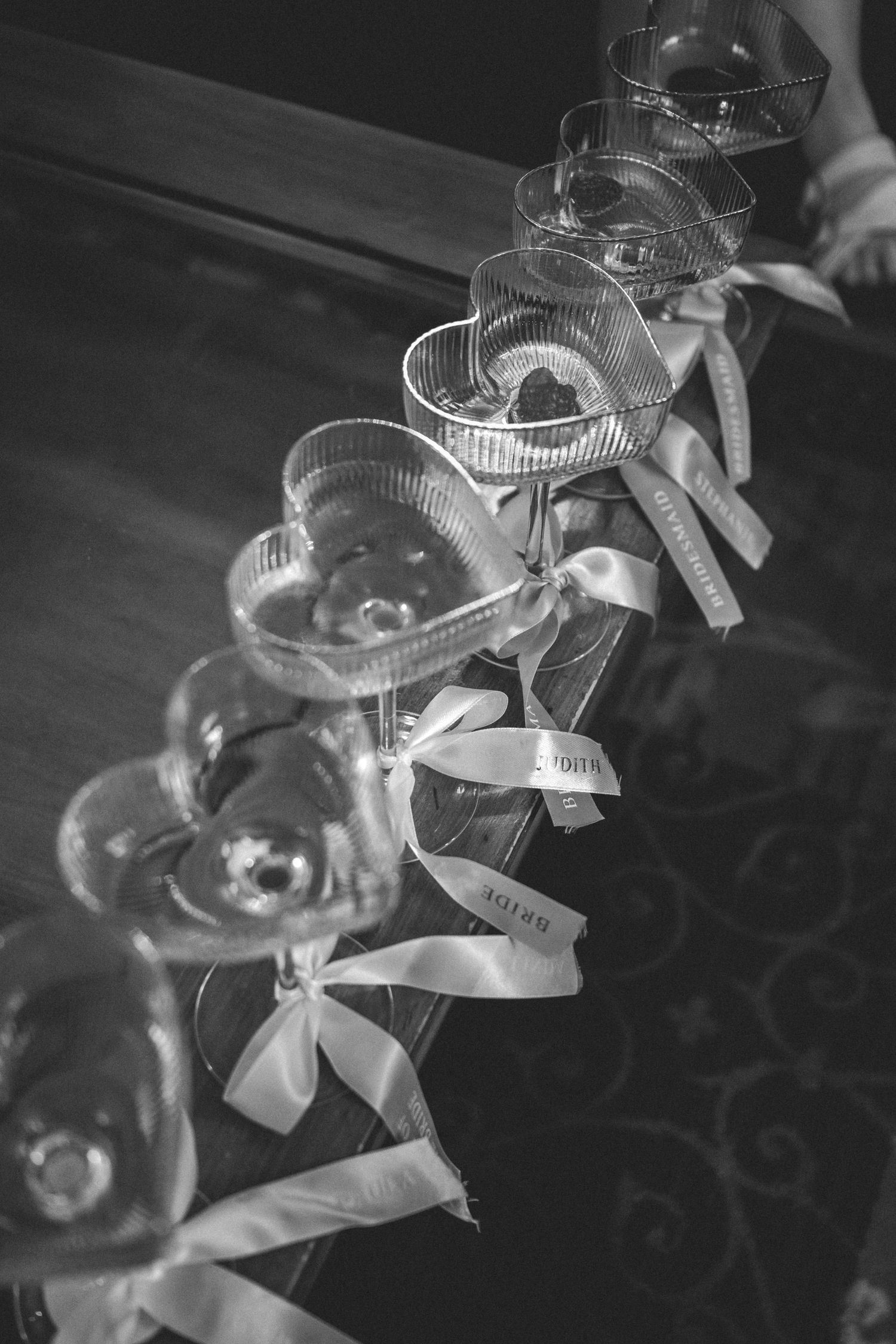 A row of heart-shaped champagne coupe glasses tied with ribbons sits on a table in a black and white wedding detail photograph.