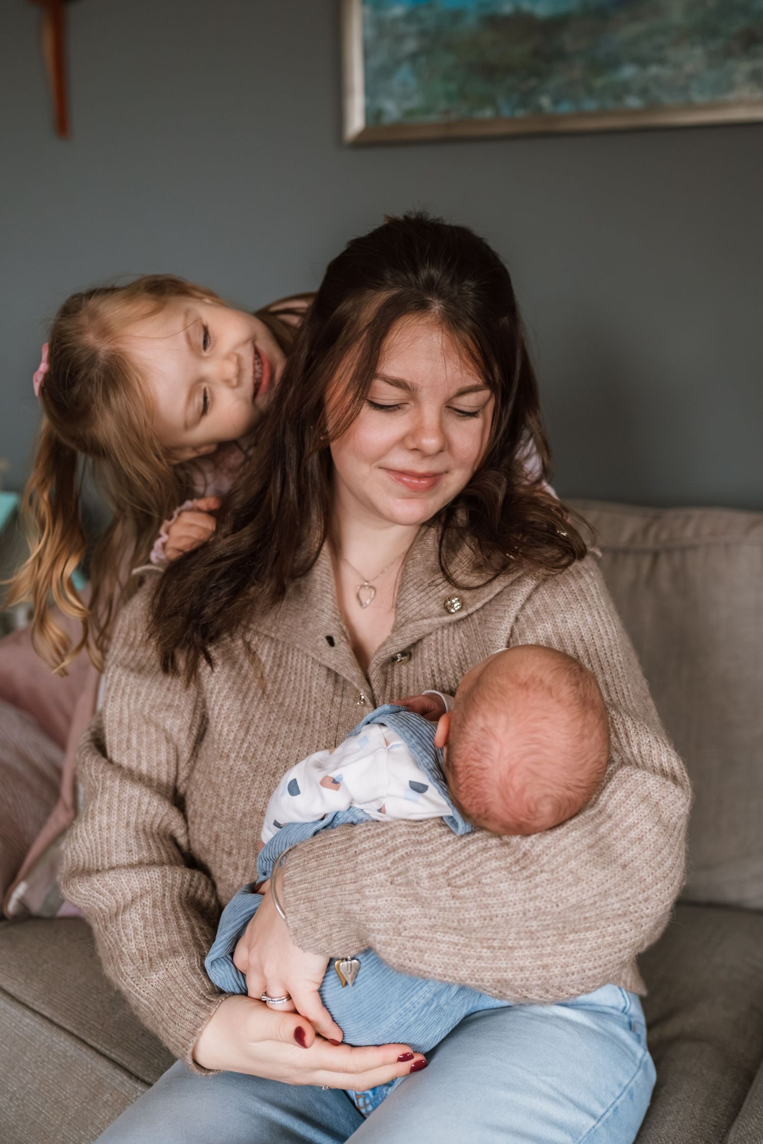 A smiling mother in a tan cardigan cradles a sleeping newborn on a cozy sofa while a young girl leans affectionately over her shoulder.