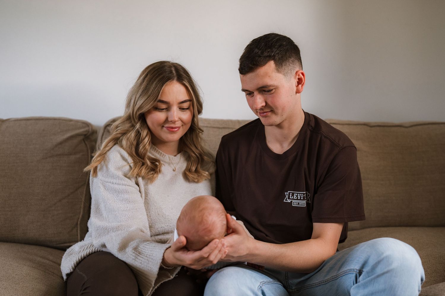 A smiling woman and a man sit together on a beige sofa, gently holding a sleeping newborn baby in their hands.