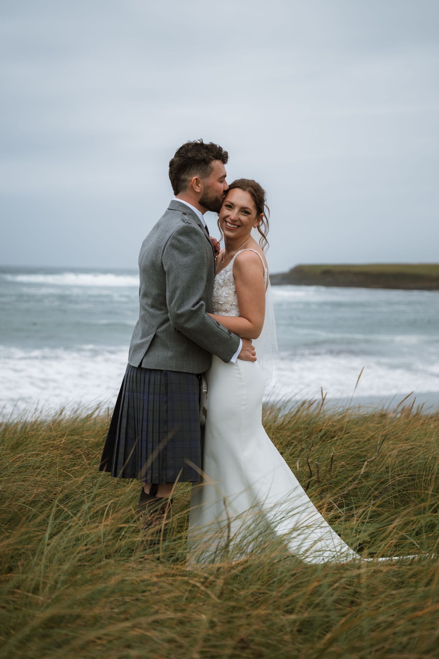 Bride in a white gown and groom in a grey jacket and kilt embrace in windswept coastal grass with ocean waves behind them.