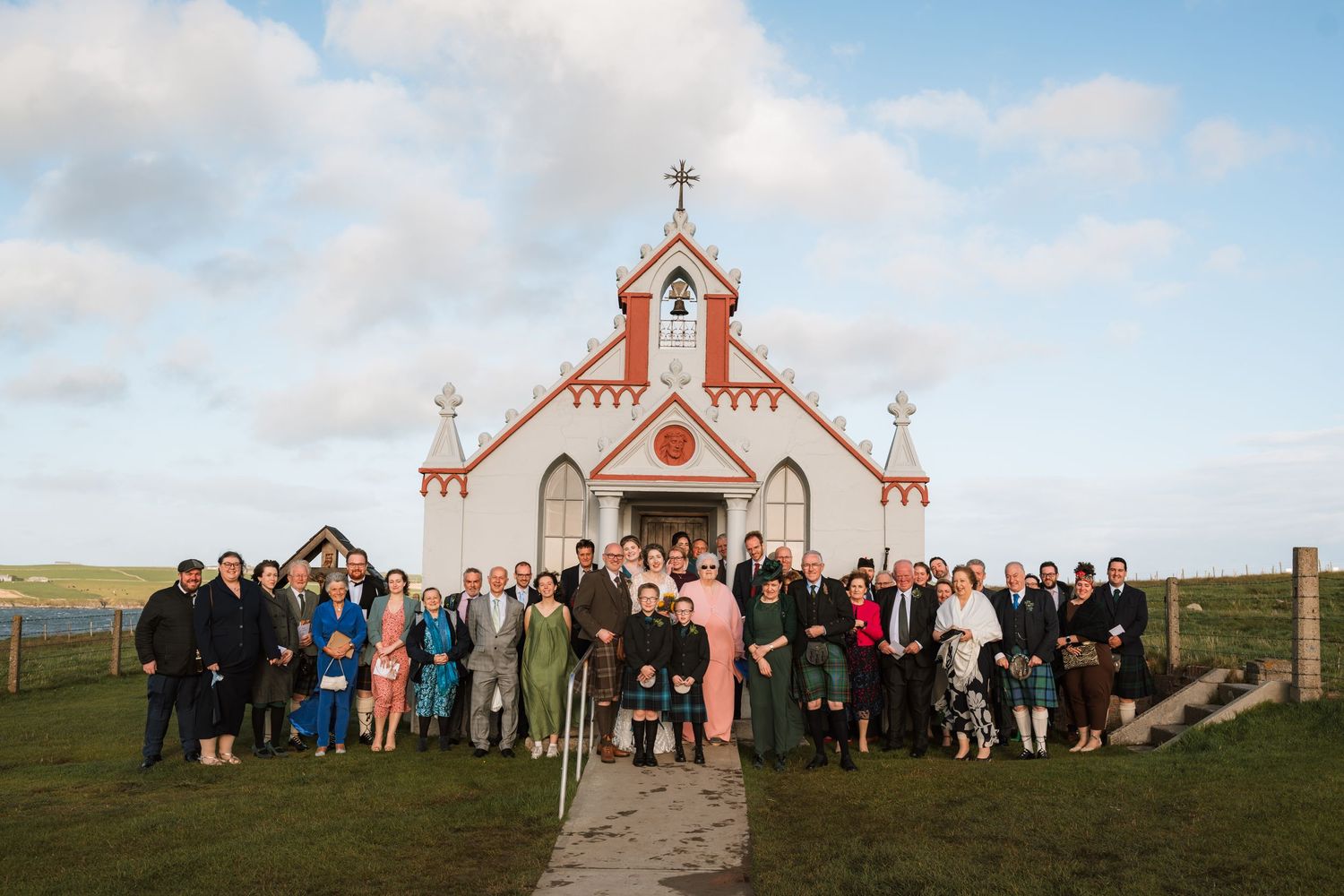 Large wedding group stands on the grass in front of a small white and red chapel under a soft blue and pink evening sky.