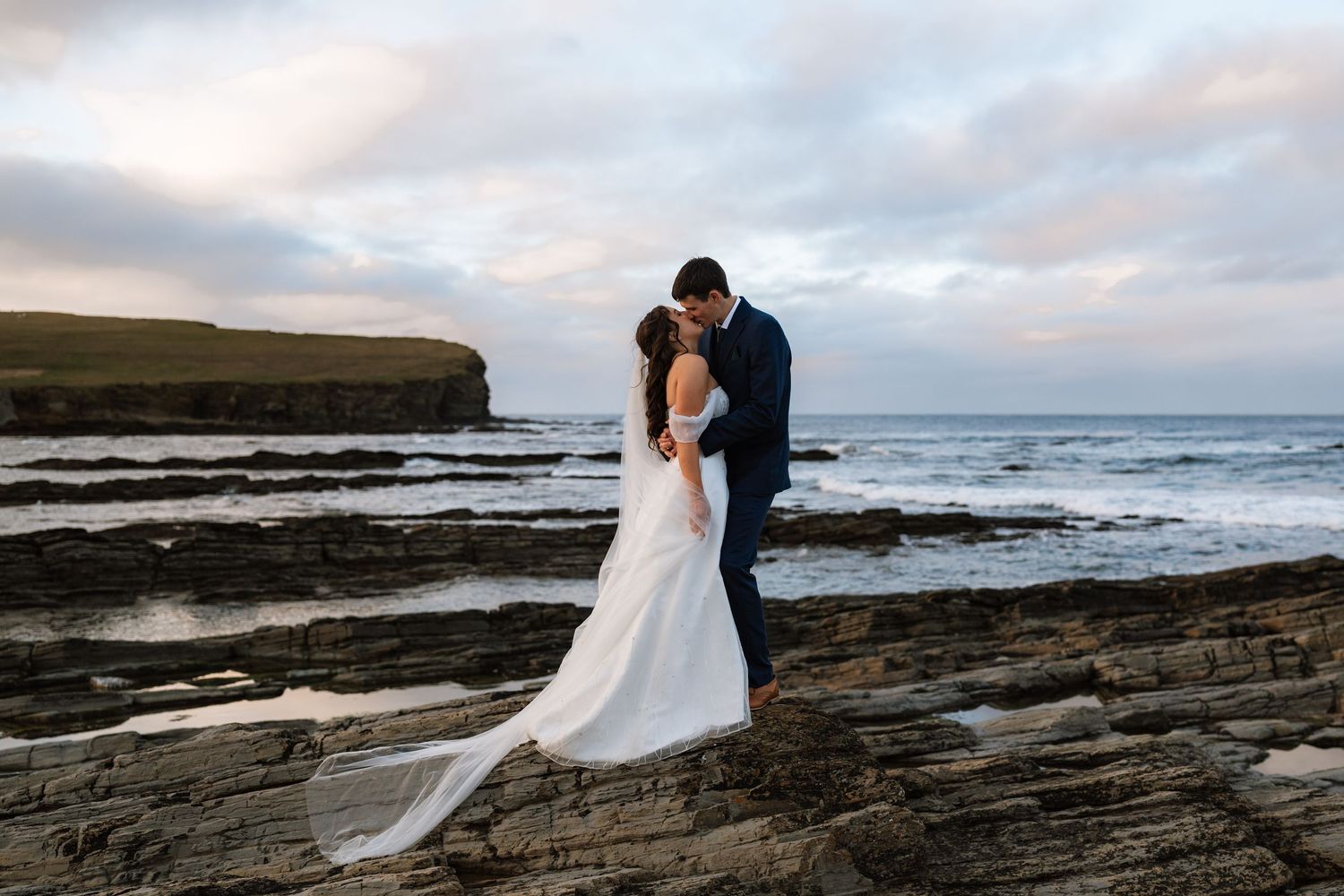 Bride and groom kiss on layered coastal rocks as waves roll in beneath a soft pastel sunset sky.