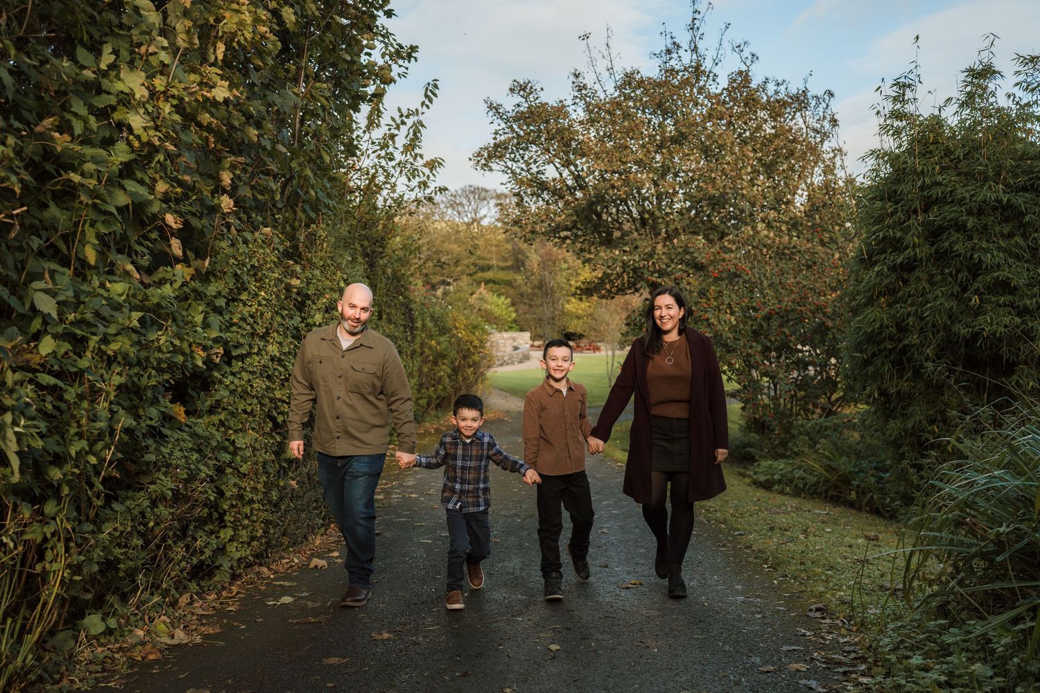 Family of four holding hands and walking down a paved path lined with green hedges and autumn trees during an outdoor family photo session.