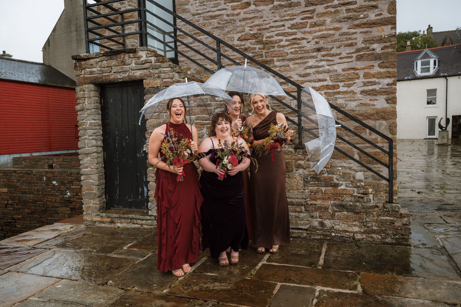 Bridesmaids in burgundy and dark dresses hold clear umbrellas and bouquets, laughing in the rain outside a stone building.
