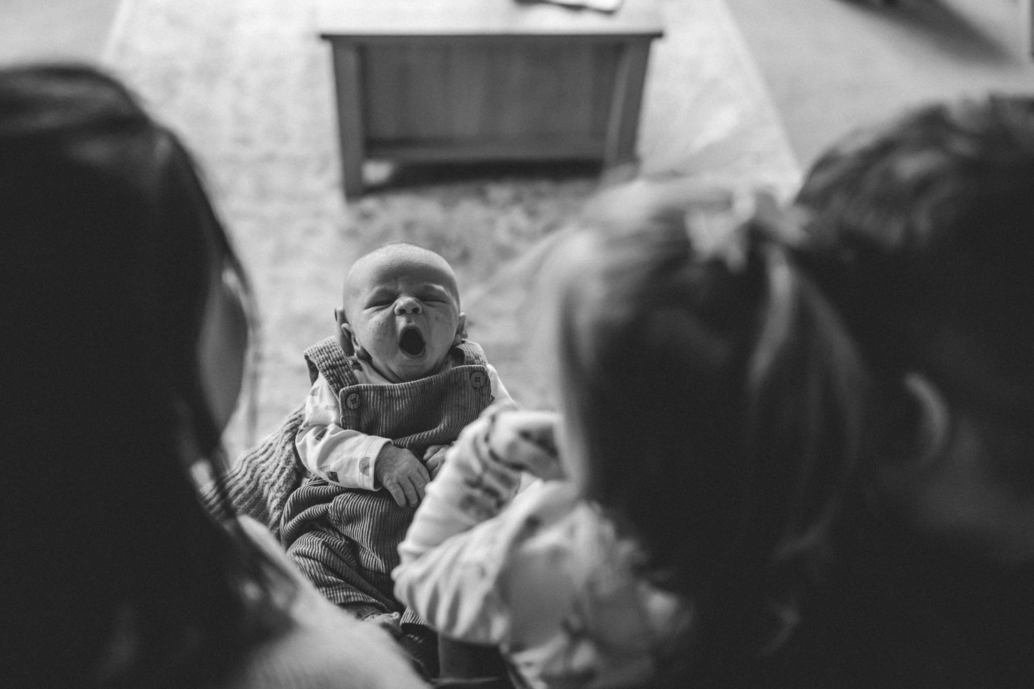 Black and white photo of a newborn baby yawning while two blurred family members lean in close over the infant.