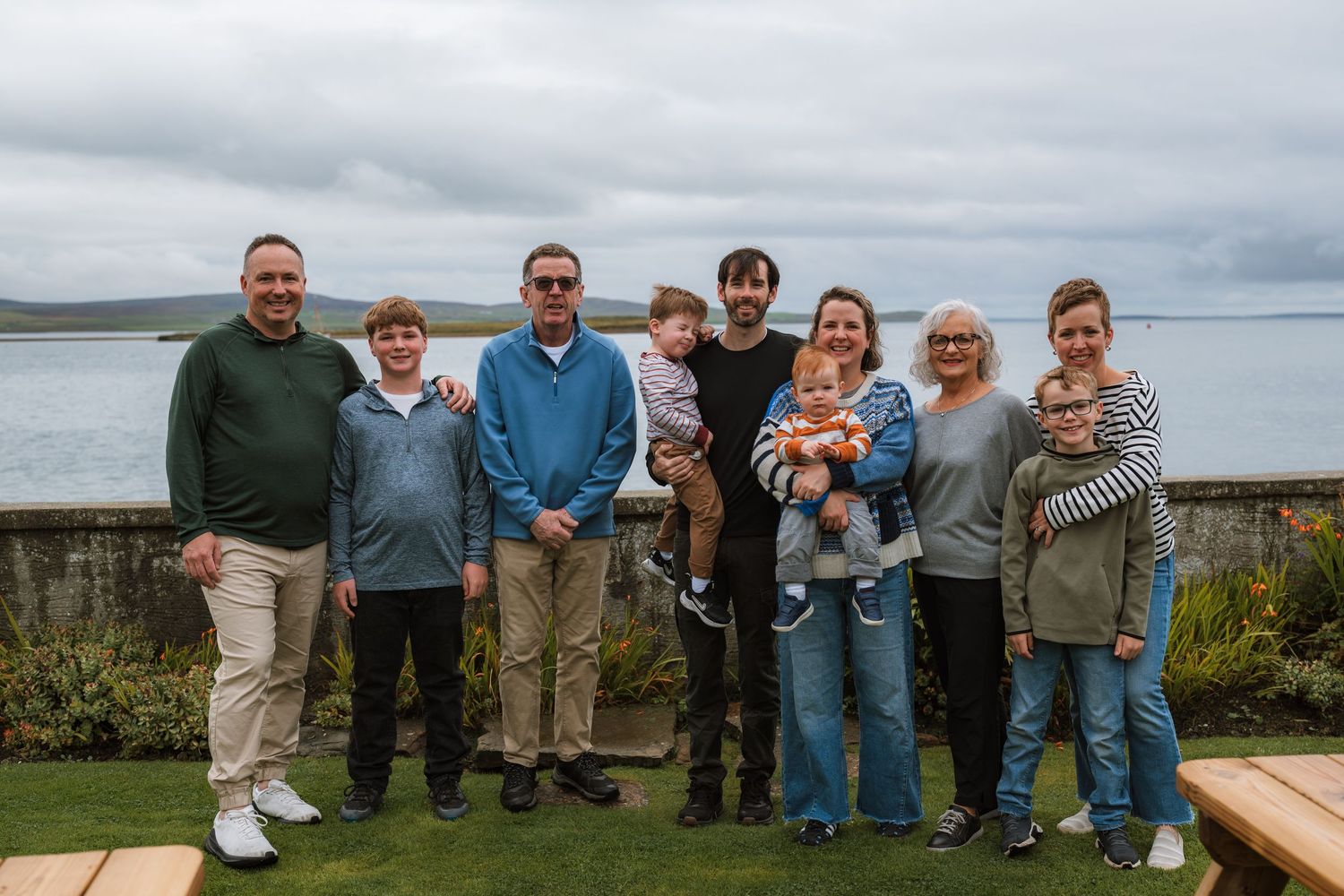 A large family of adults and children poses together on a lawn beside a stone wall, with a calm bay and hills behind.