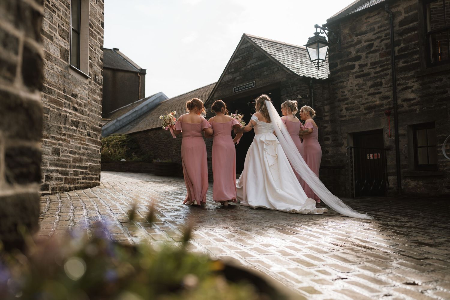 Bride in a long veil walks a cobblestone lane with four bridesmaids in blush pink dresses.