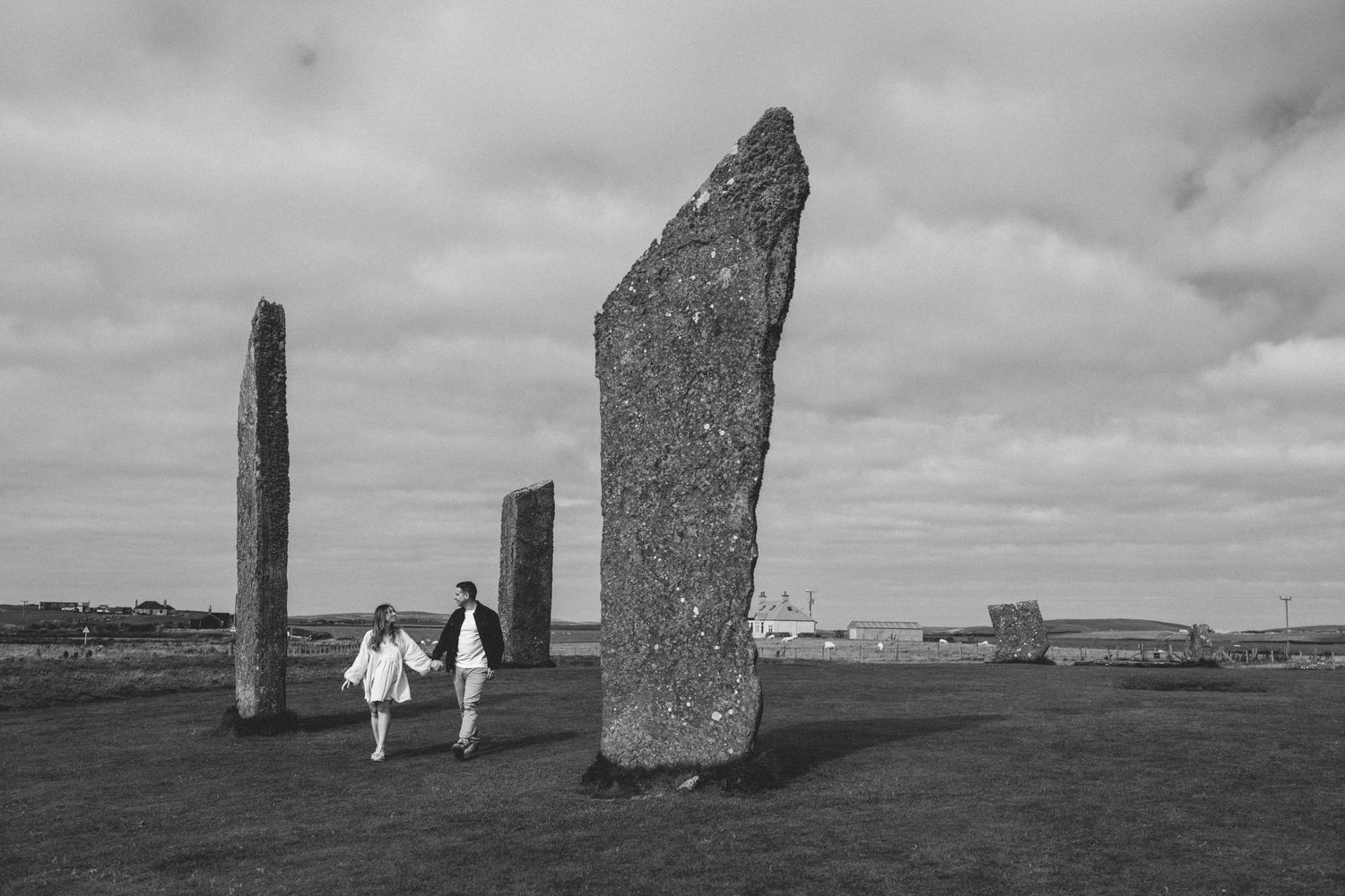 Couple walks hand in hand among ancient standing stones under a cloudy sky in black and white.