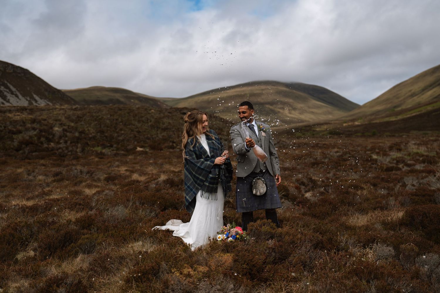 Bride and groom in tartan wraps and kilt stand together on a wild Scottish moor surrounded by rolling mountains and heather.