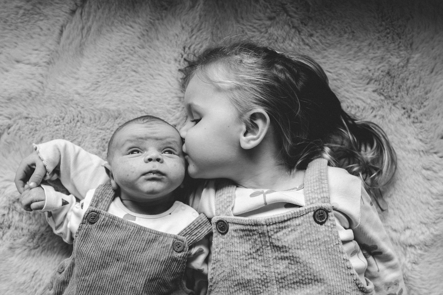 Black and white photo of a young girl in denim overalls kissing the cheek of a newborn baby lying beside her on a soft blanket.