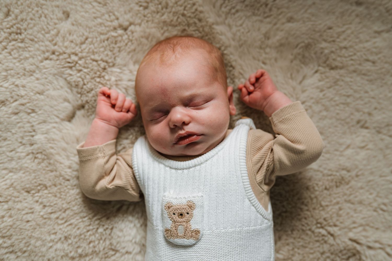 Sleeping newborn baby with arms raised, wearing a cream vest with a teddy bear patch, resting on a soft fluffy sheepskin rug.