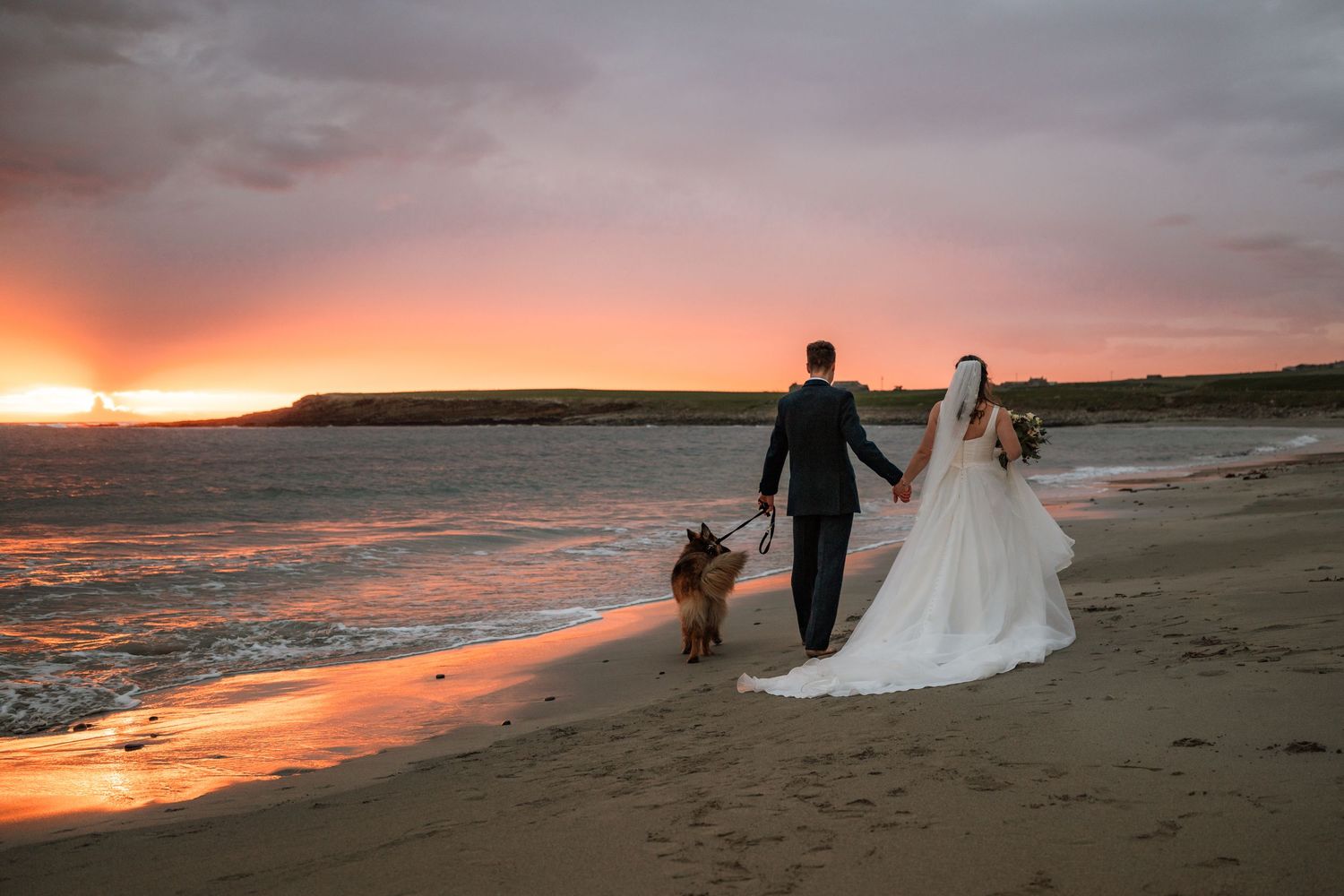 Bride and groom walk hand in hand with their dog along a beach at sunset beneath a vivid pink and orange sky.