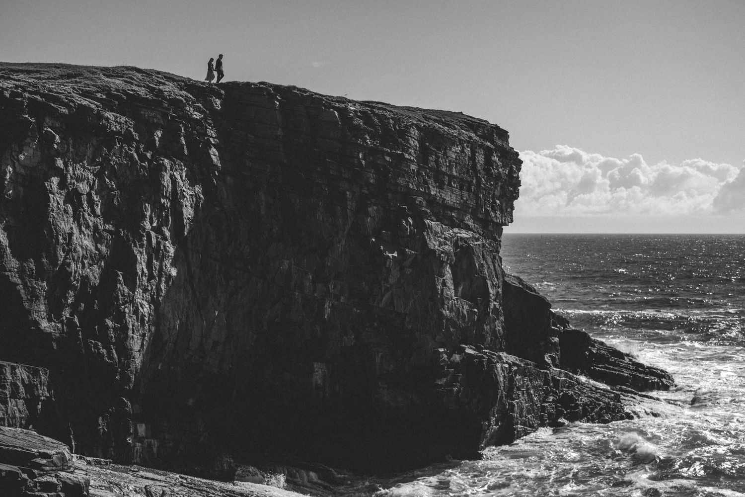 A black and white view of a couple standing on top of a huge rugged sea cliff above crashing ocean waves.