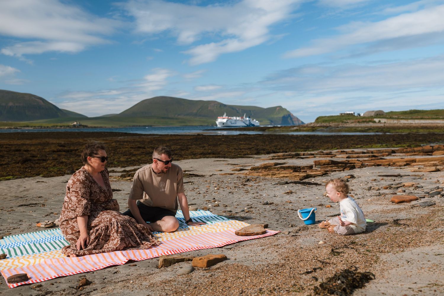 Two adults relax on a striped picnic blanket on a rocky beach while a toddler plays with a bucket near a distant ferry.