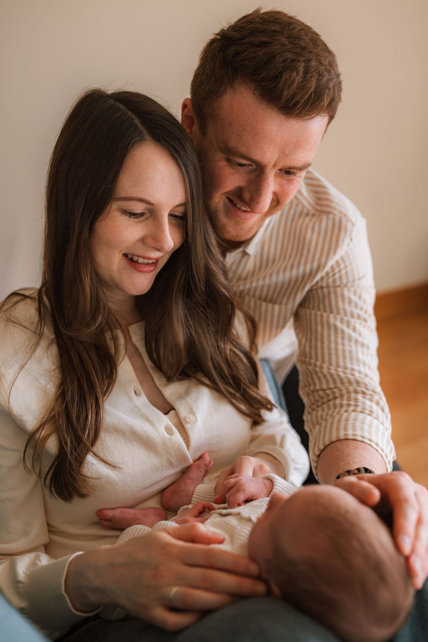 Smiling couple in cream outfits sit close together, gazing lovingly down at their sleeping newborn baby in a bright, cozy home.