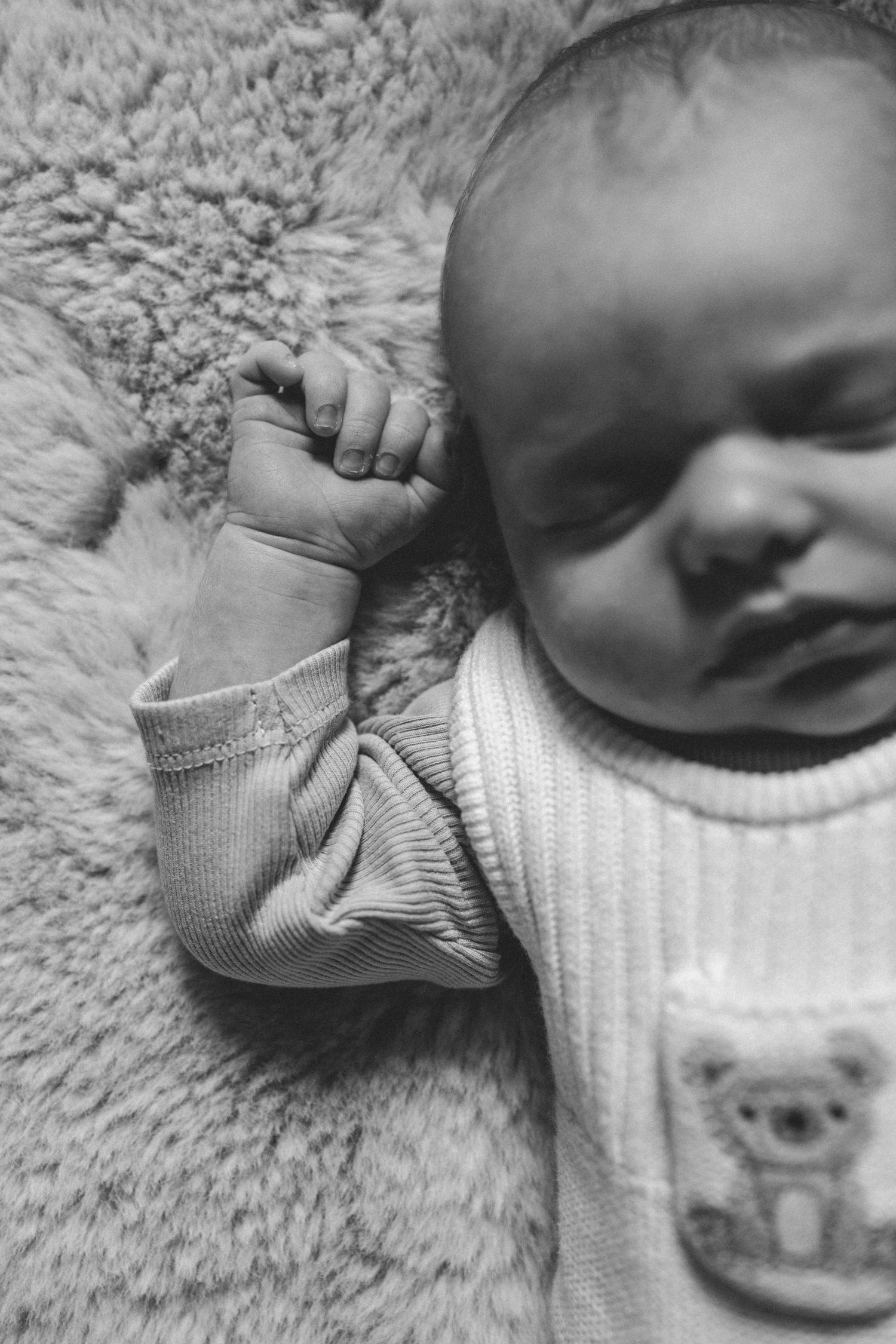 Black and white close-up of a sleeping newborn baby with a tiny fist raised, wearing a ribbed sweater with a koala bear pocket on a fluffy b