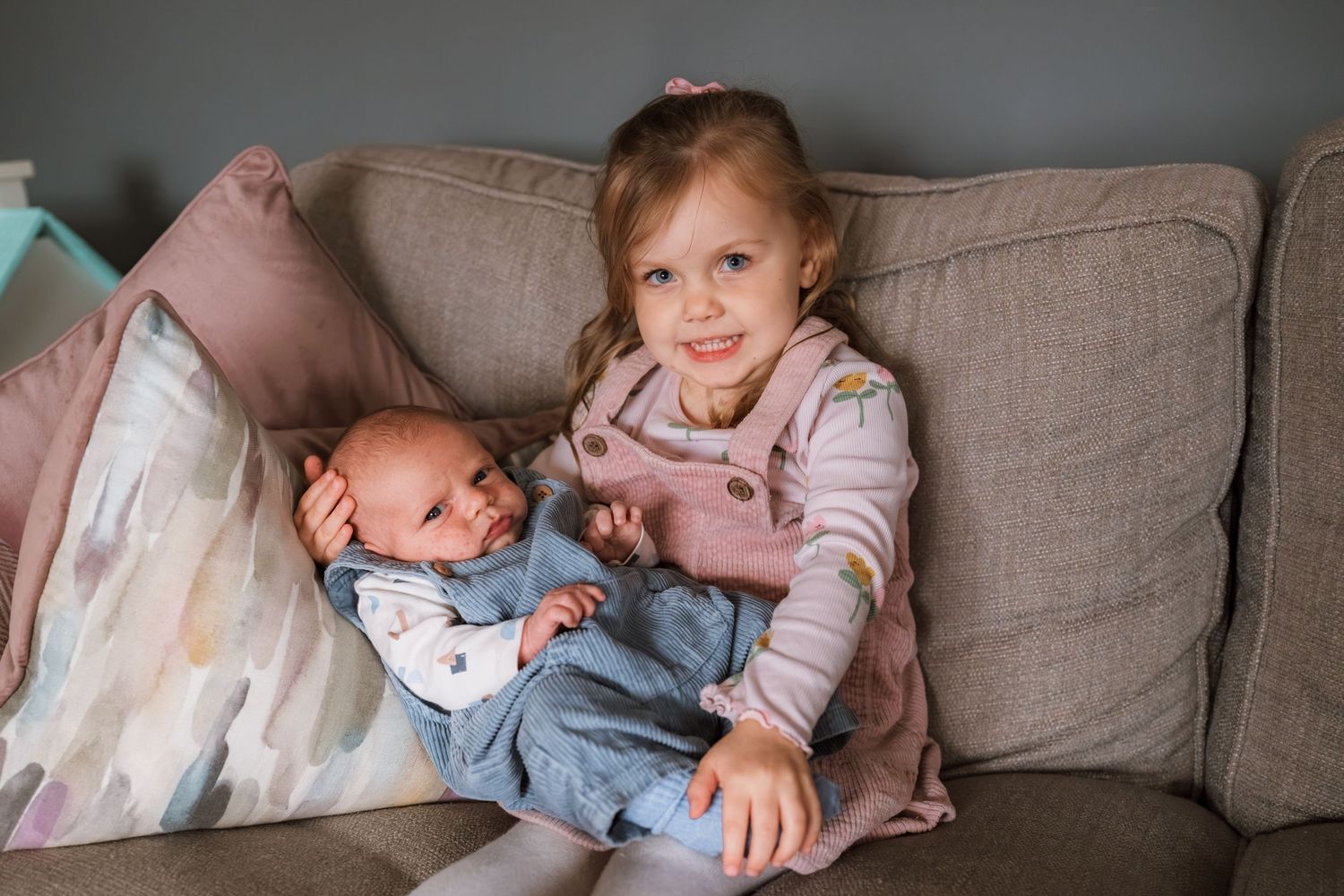 Smiling toddler girl in a pink pinafore sits on a gray sofa cradling her sleeping newborn baby brother in a blue outfit.