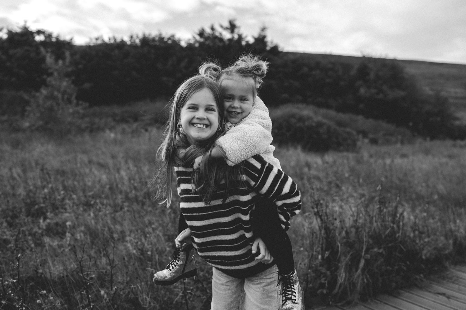 Black and white photo of a smiling girl giving a younger girl a piggyback ride in a grassy field with trees behind.
