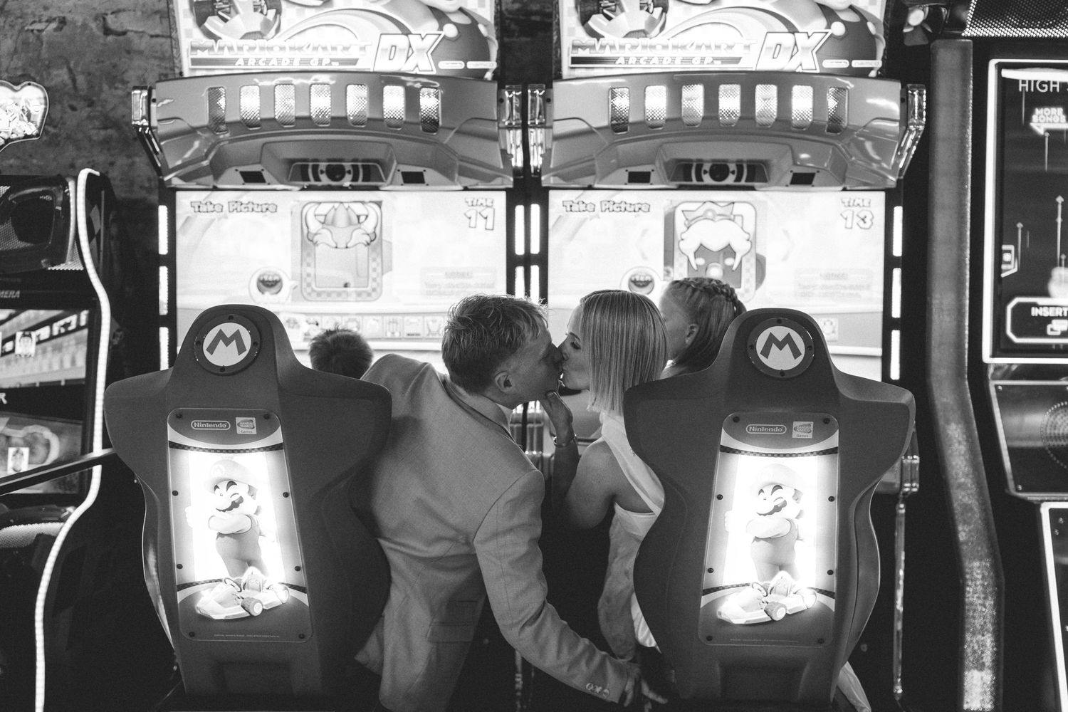 A couple shares a kiss while seated at colorful arcade racing machines in a black and white wedding photograph.